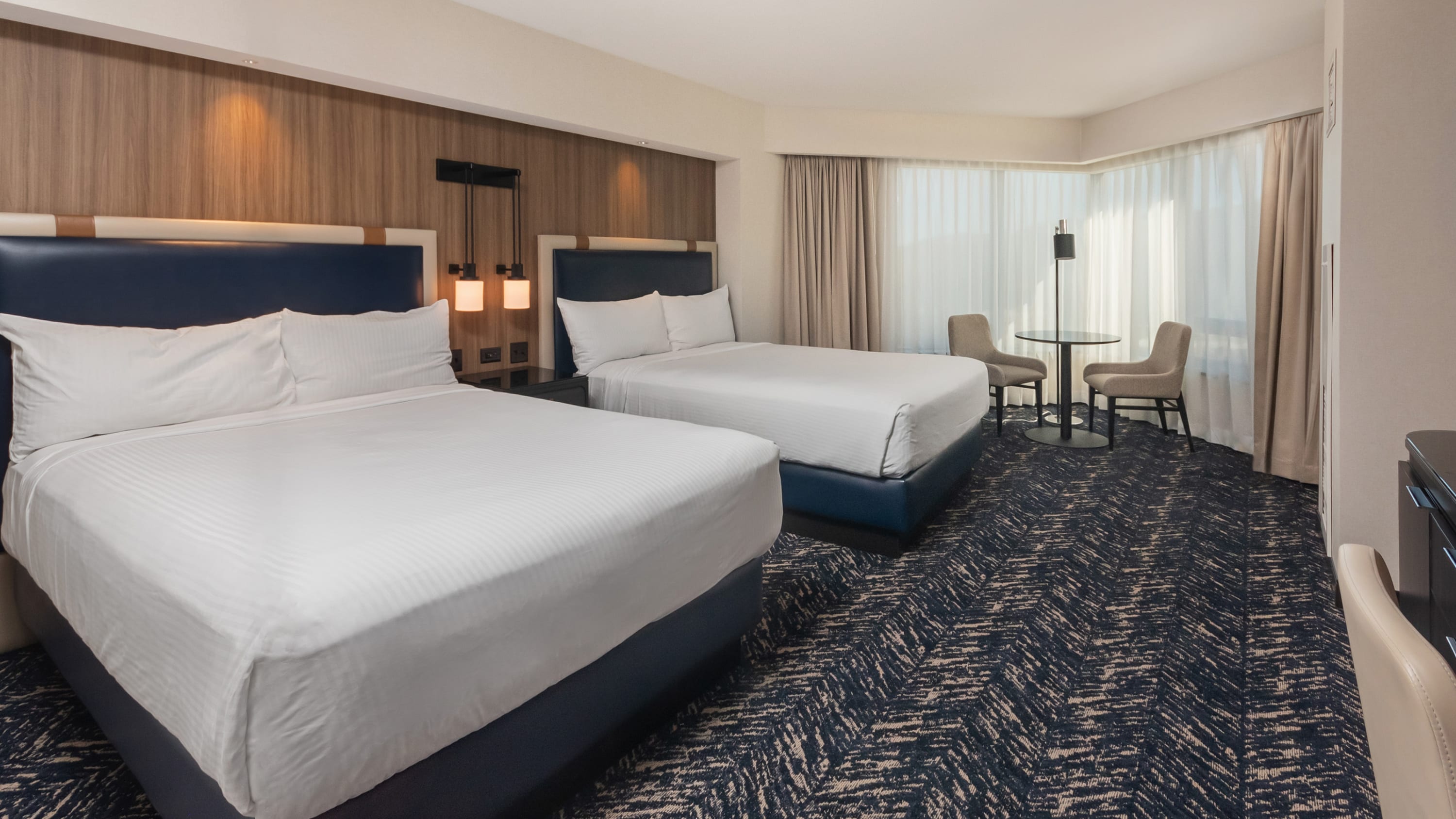 A hotel room with blue striped carpet, two beds with white sheets, a window with a table and two chairs in front of it