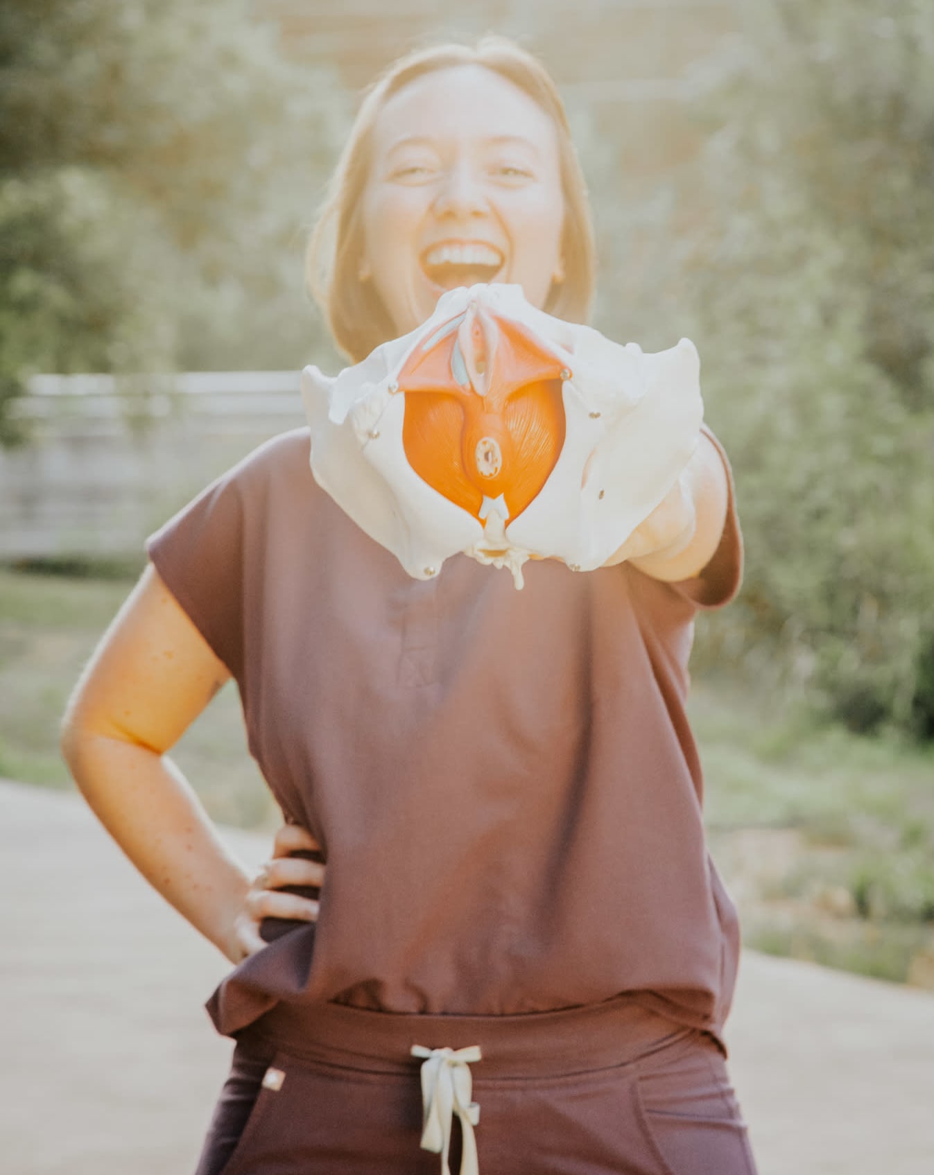 Dr. Ashe wearing purple scrubs with natural greenery in the background. She is smiling and holding a 3D pelvic model.