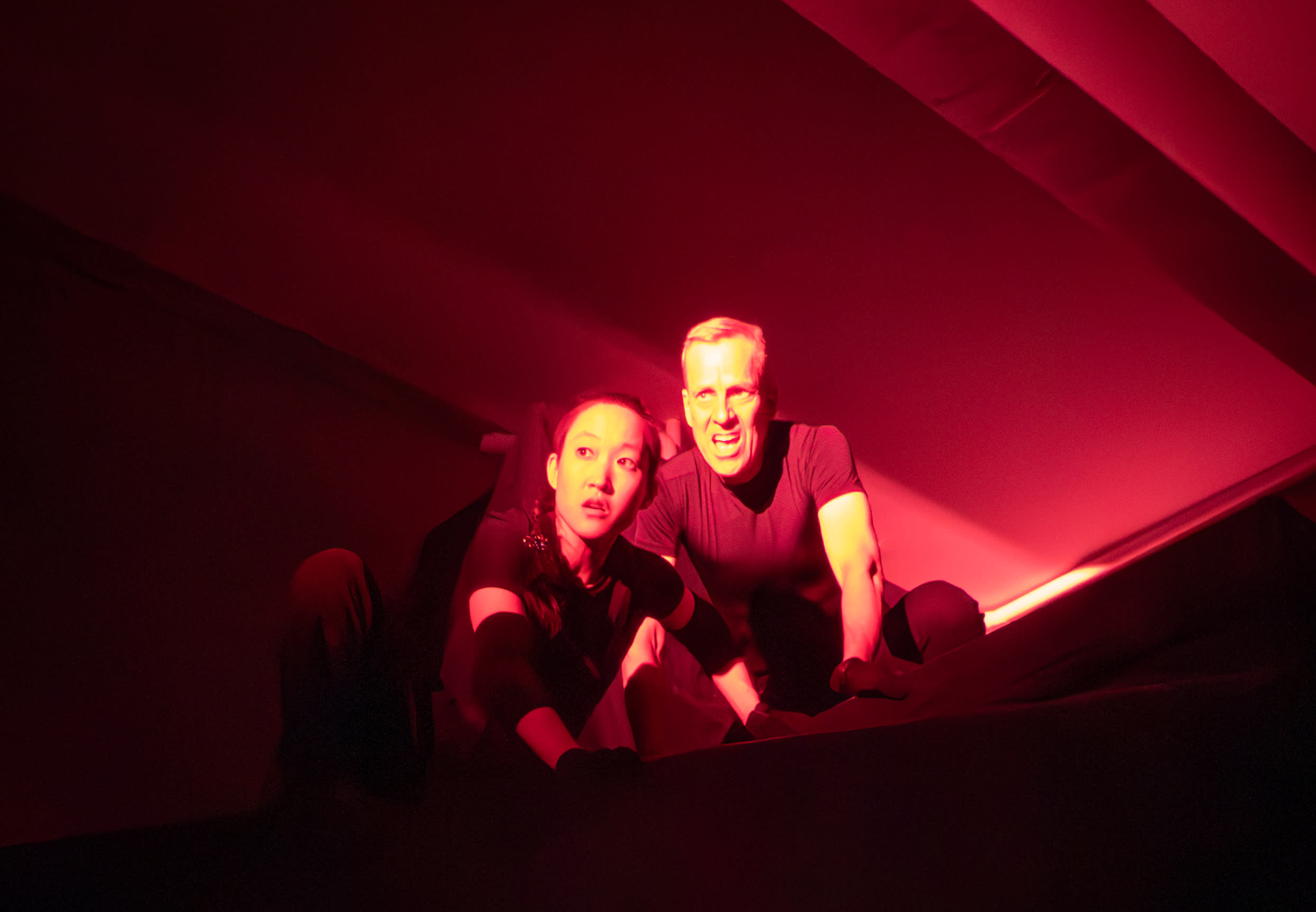 two actors in all black crouched in a small, red space