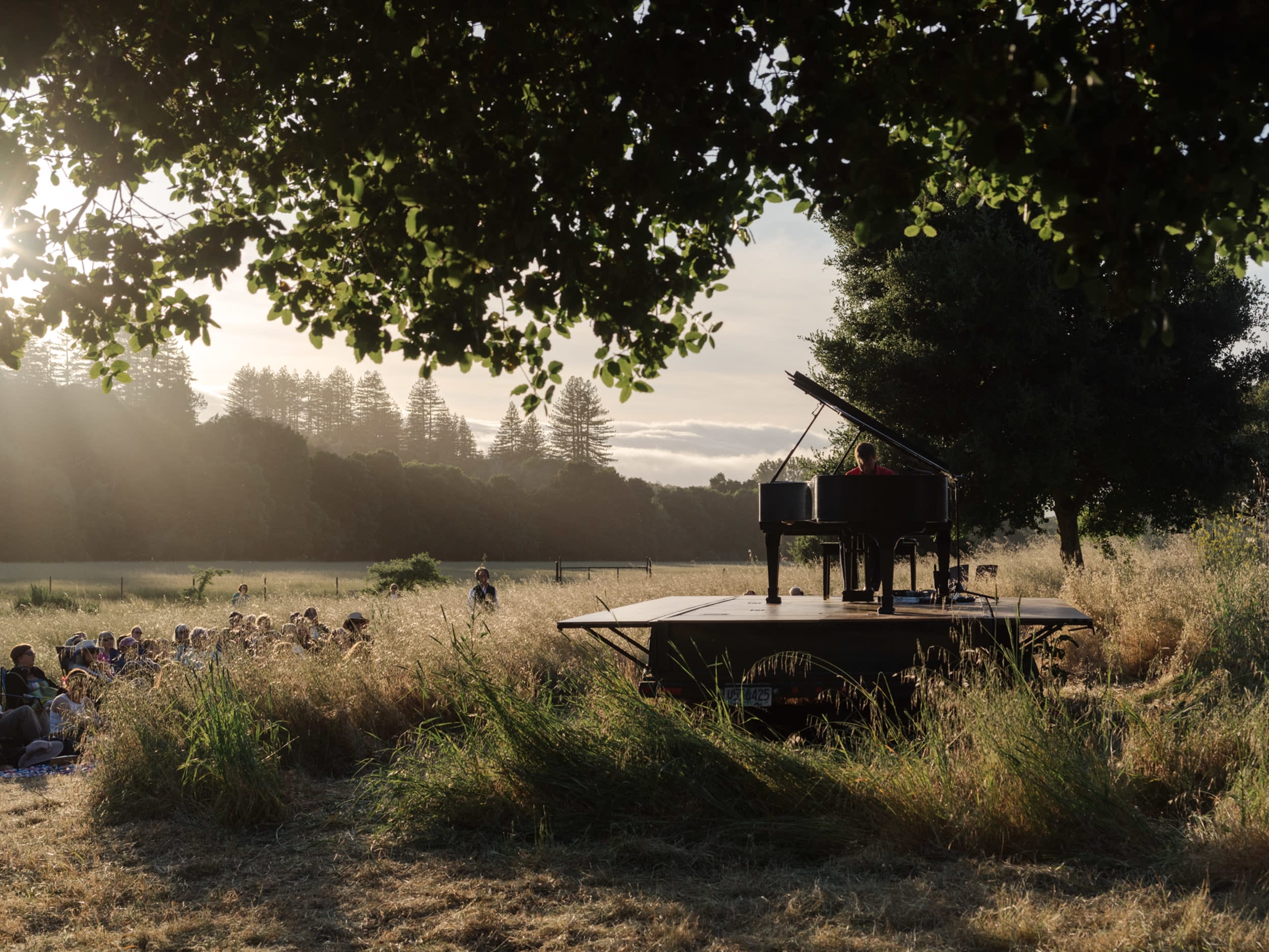 Hunter Noack sits at a piano at sunset in a lush green area surrounded by trees. Audience members are seated on the ground w