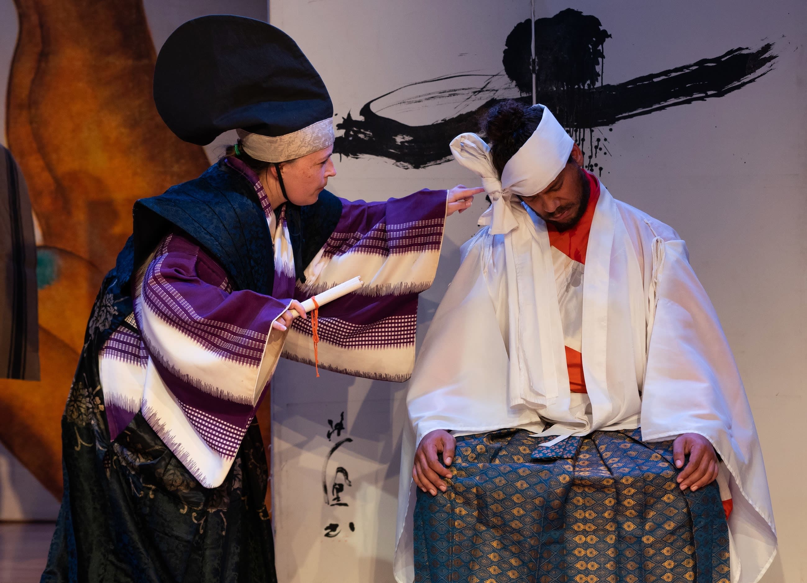 Two actors in traditional Kyogen costumes—one seated in a chair with his eyes closed, appearing to be asleep, with a white fa