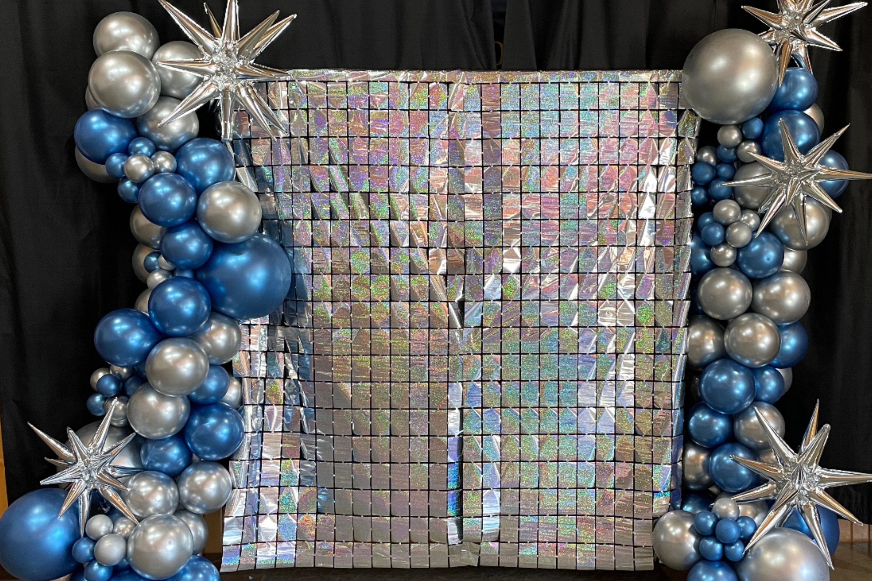 Shimmering iridescent backdrop flanked by a silver and blue balloon garland with metallic starburst balloons.