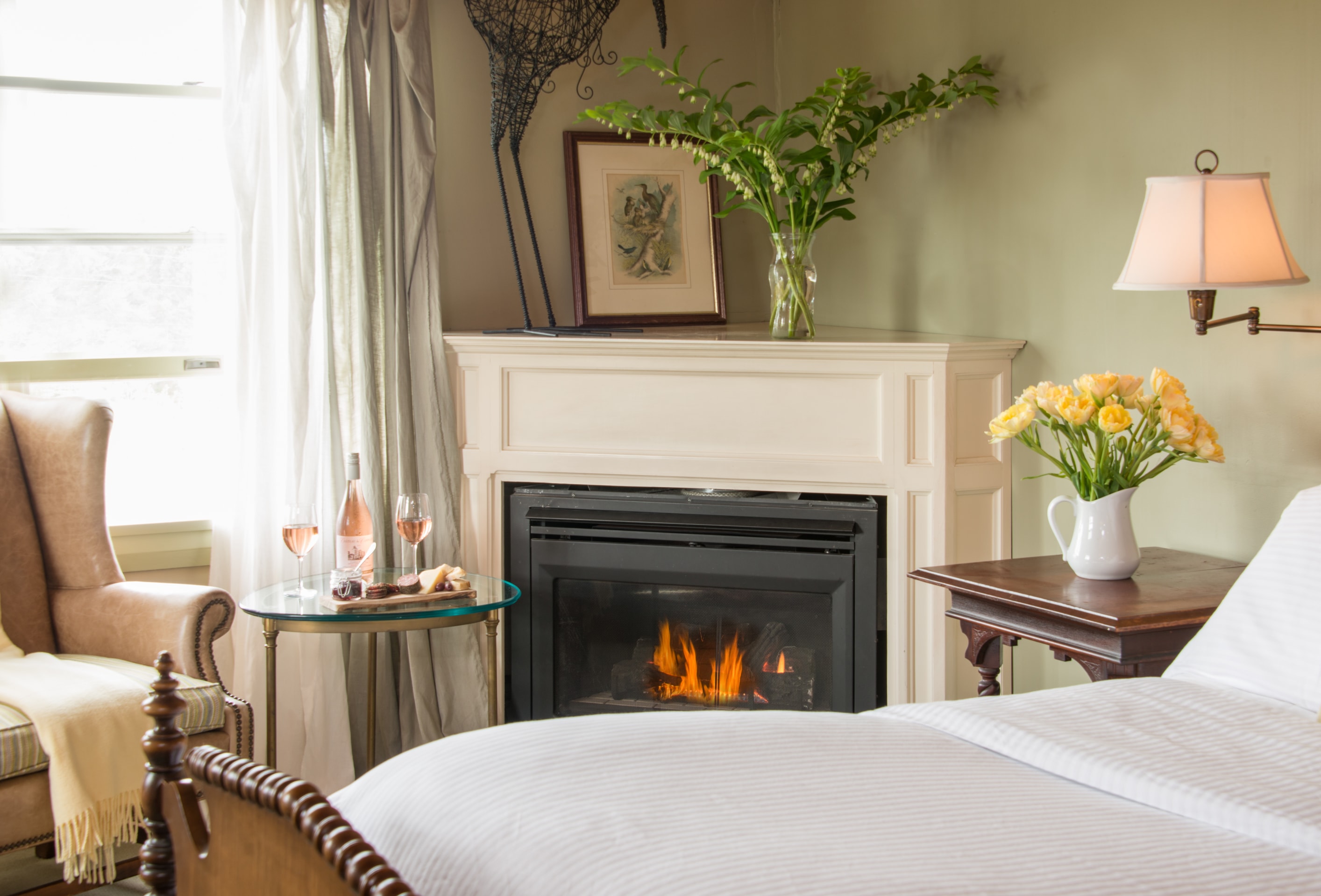 A guest room with soothing green walls, a fire in the corner fireplace, flowers in vases, a window to the back yard.