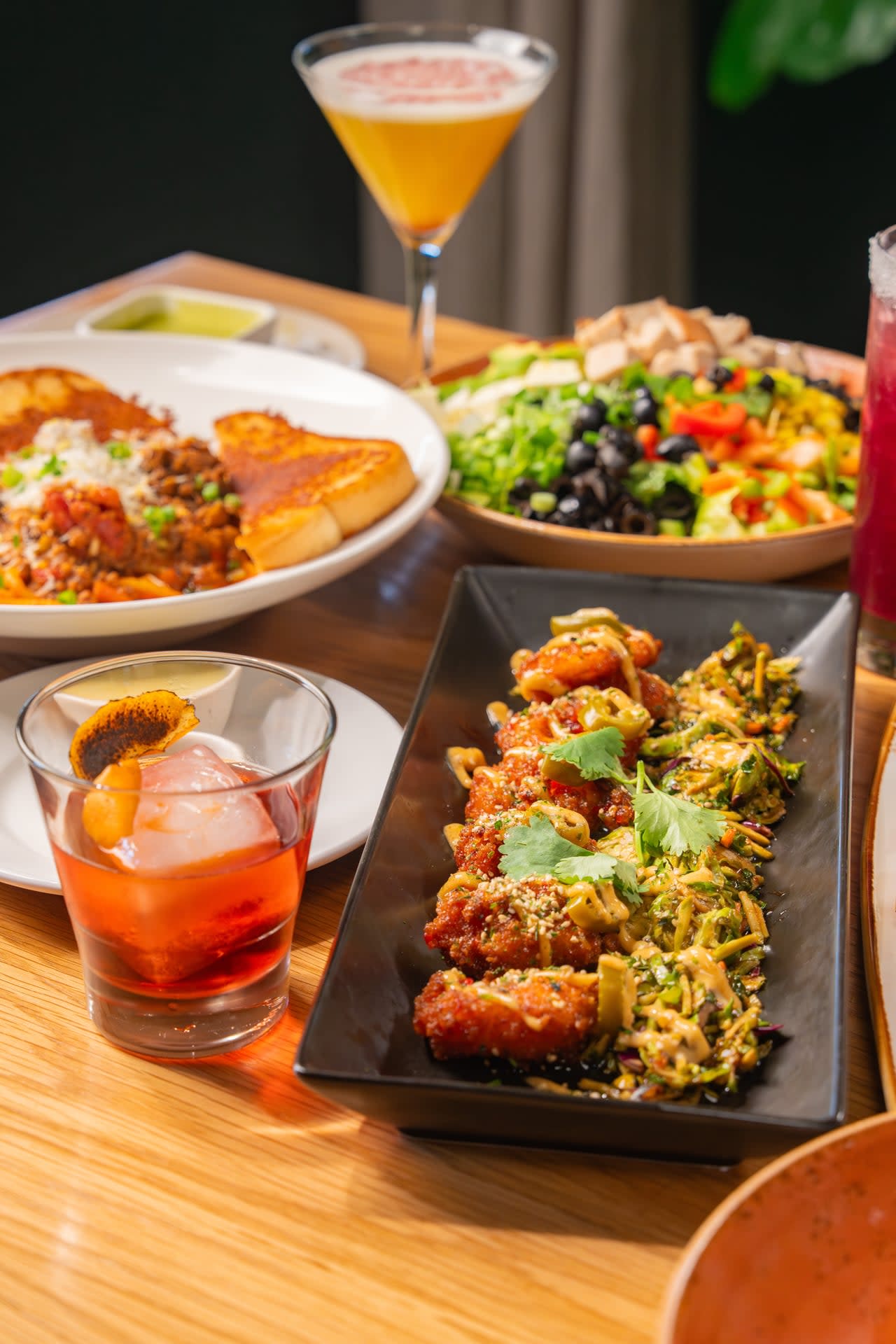A spread of Rascal Modern Diner dishes including crispy chicken, pasta, a chopped salad, and craft cocktails in