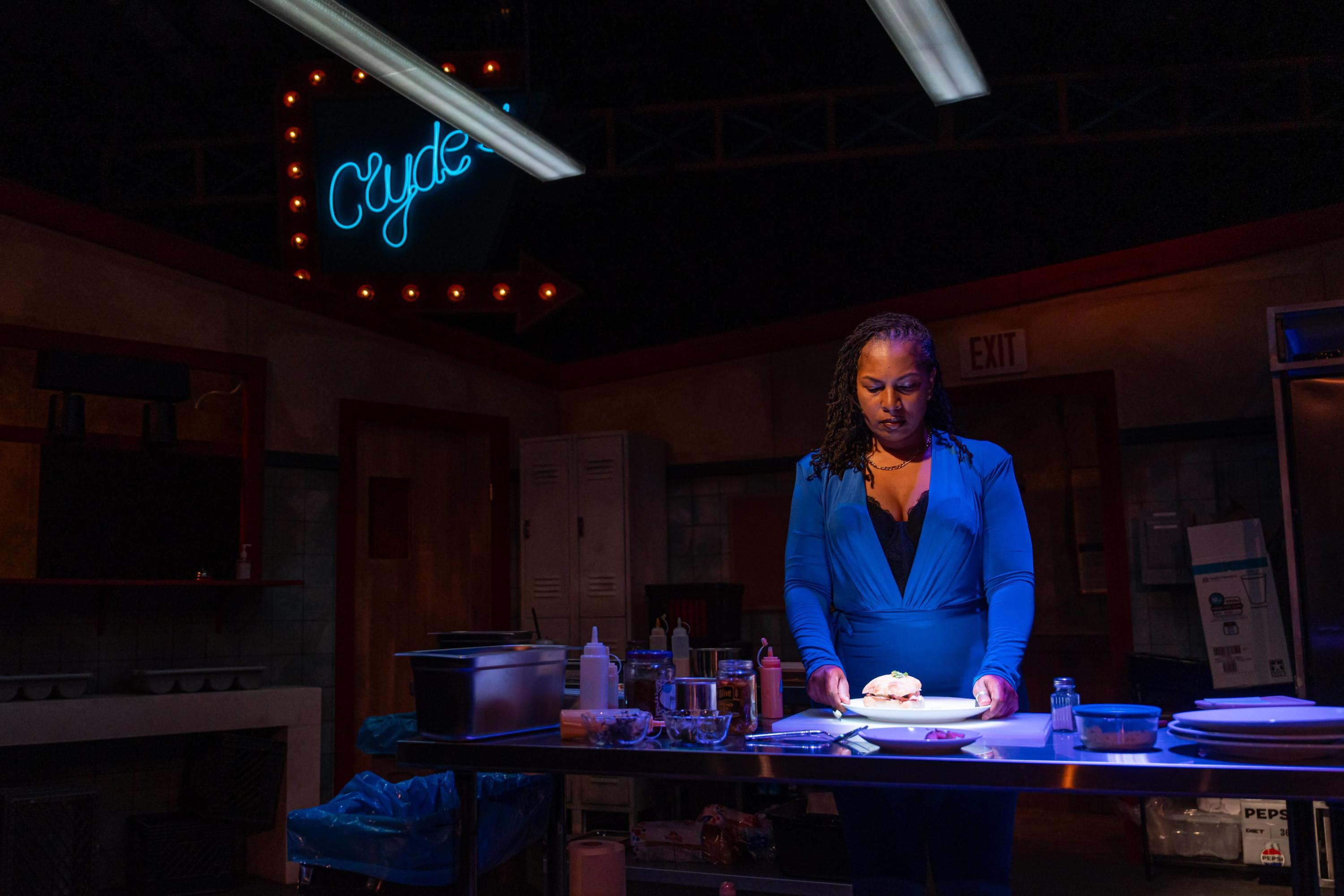 Black female actor in blue looks at a sandwich on a plate