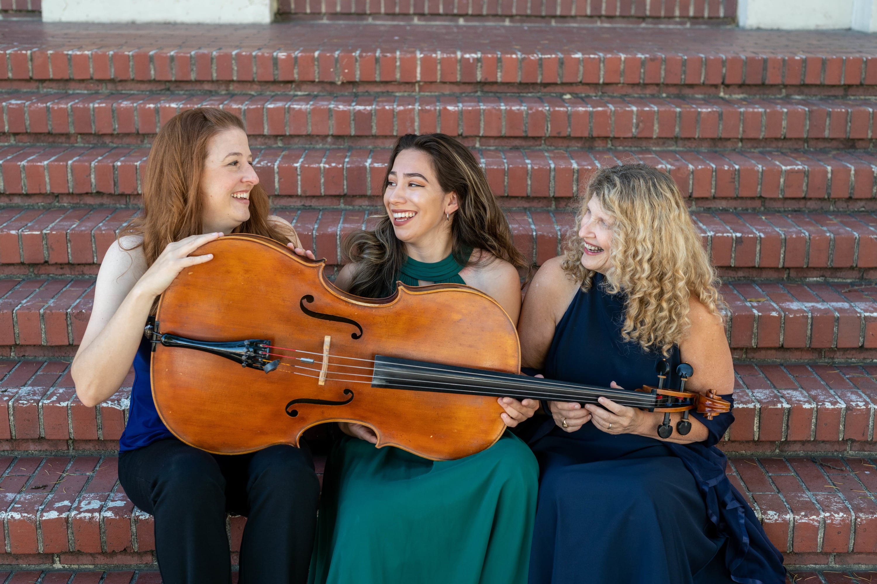 L to R Margaret Halbig, Nanette McGuinness, Abigail Monroe sitting on red brick steps, holding cello horizontally