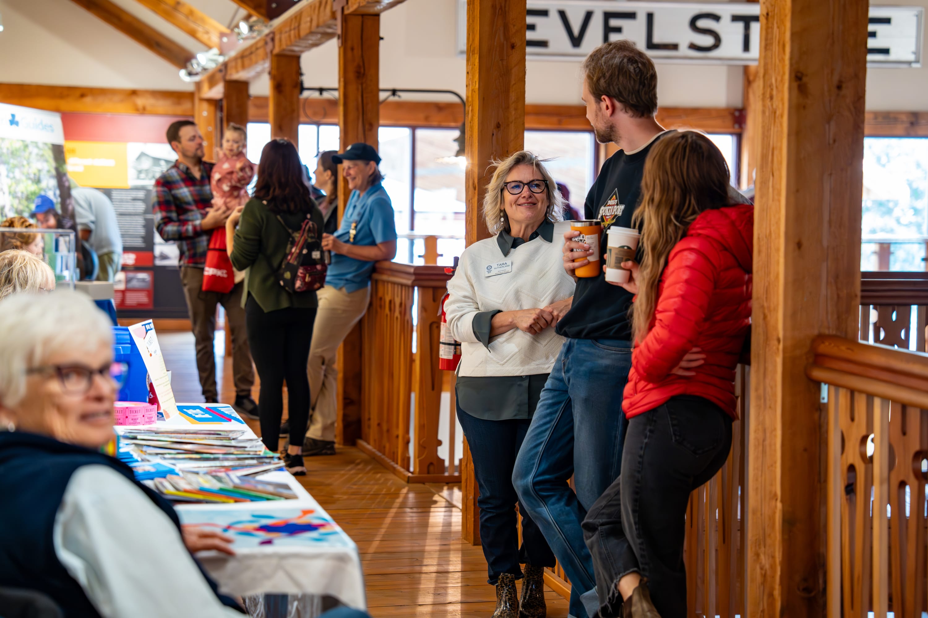 In focus are three people standing holding coffees smiling while leaning against the railing of the mezzanine.