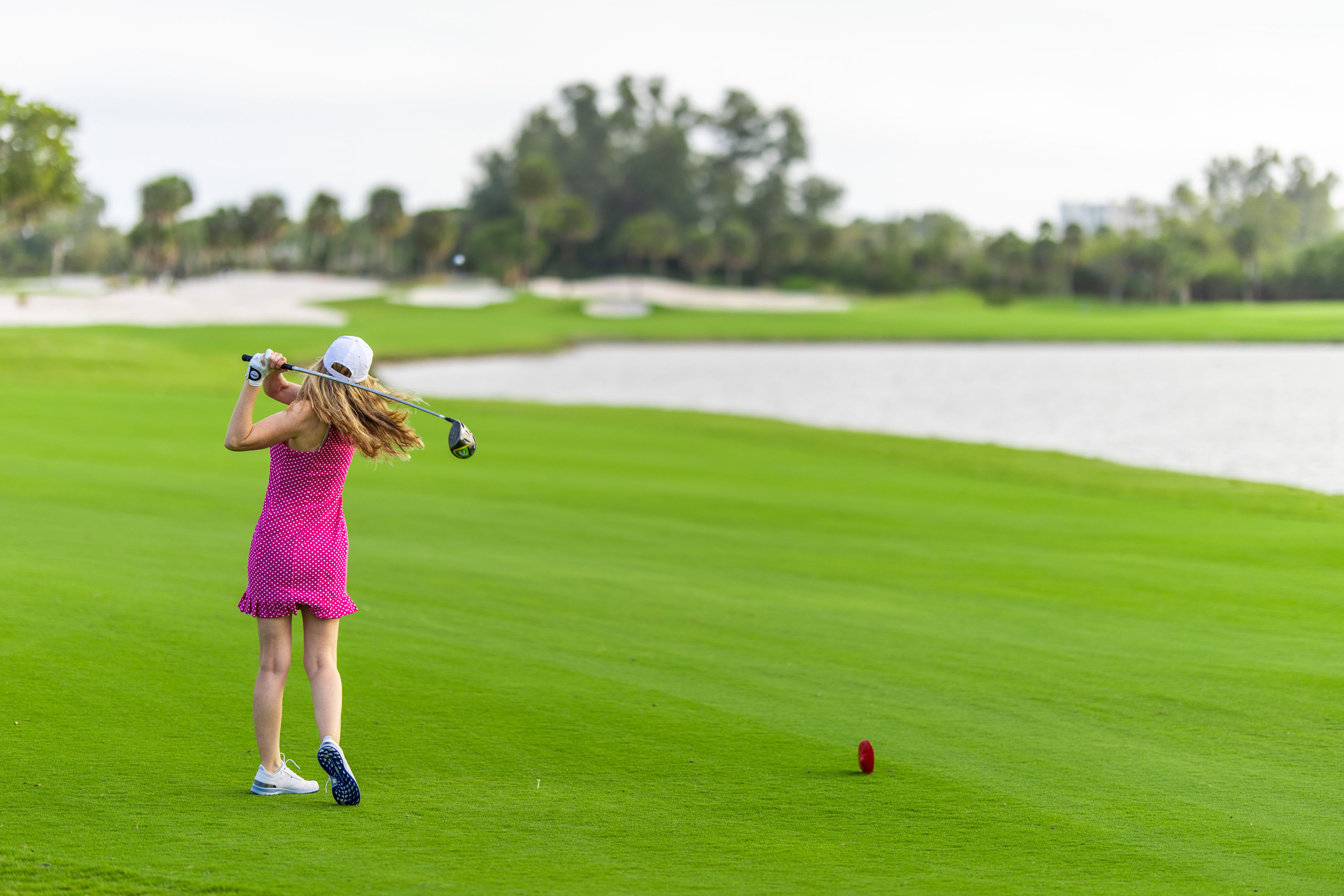 Lady in pink dress hitting swinging a golf club on a cloudy day on green golf course