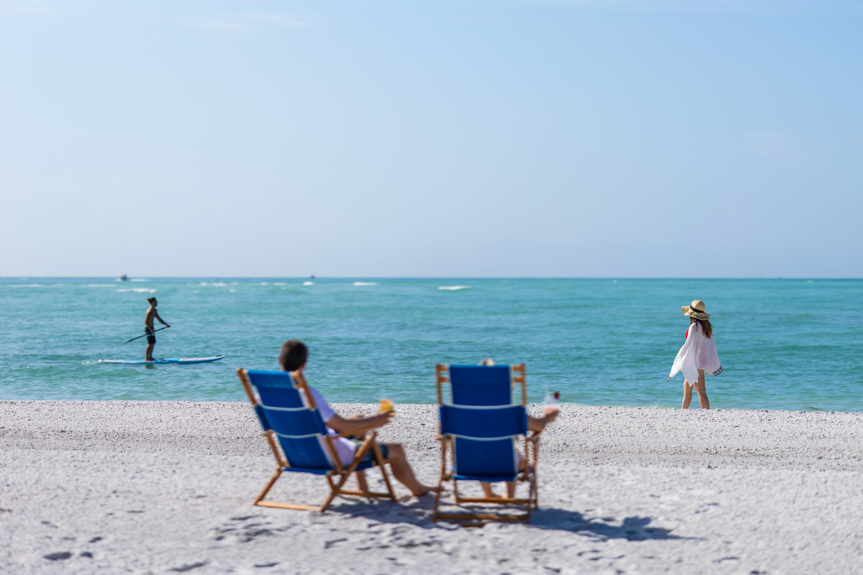Boat in the distance, man on paddleboard, a couple in blue beach chairs and woman walking on the beach