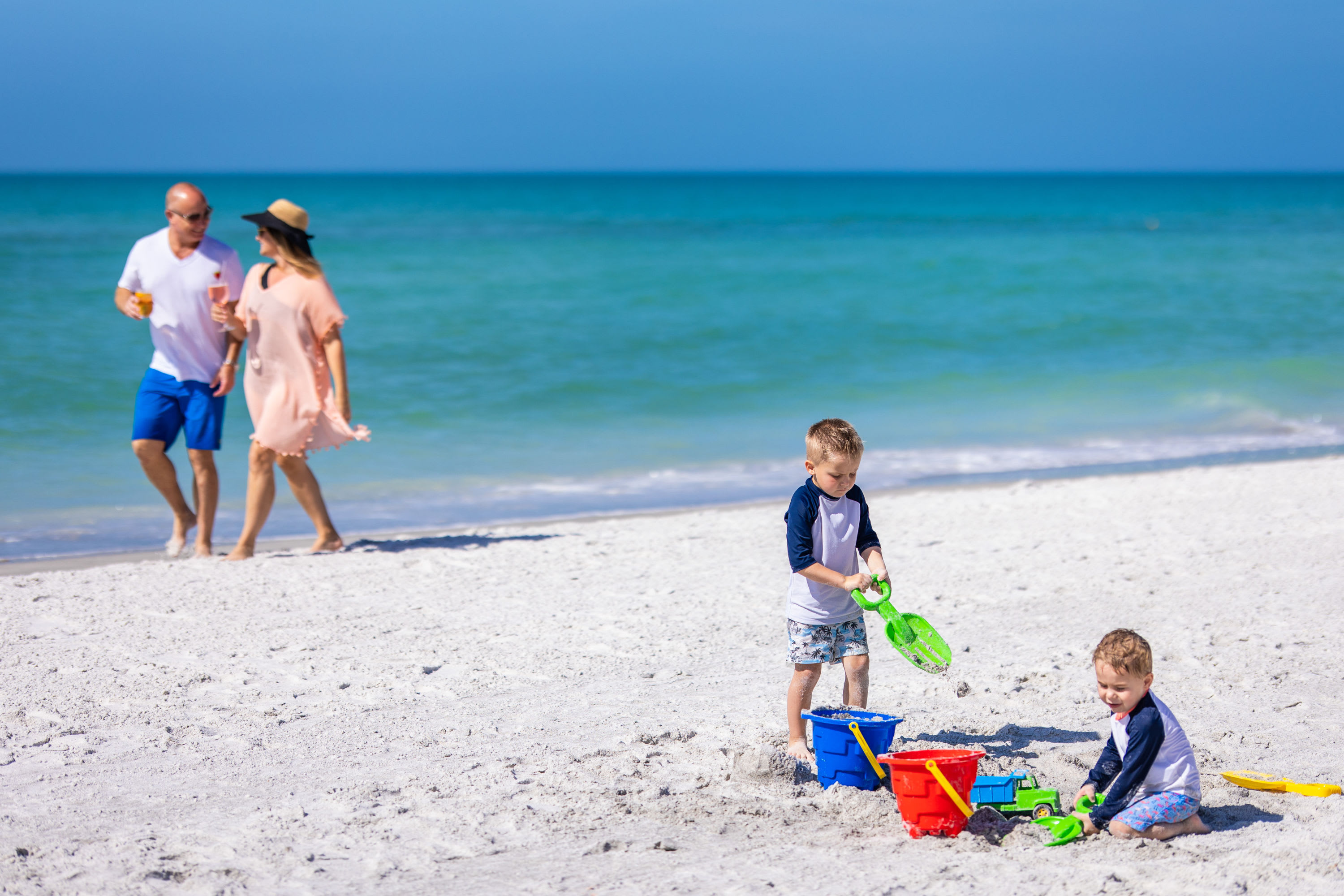 Kid's playing with sand toys on the beach as couple holding cocktails walk by