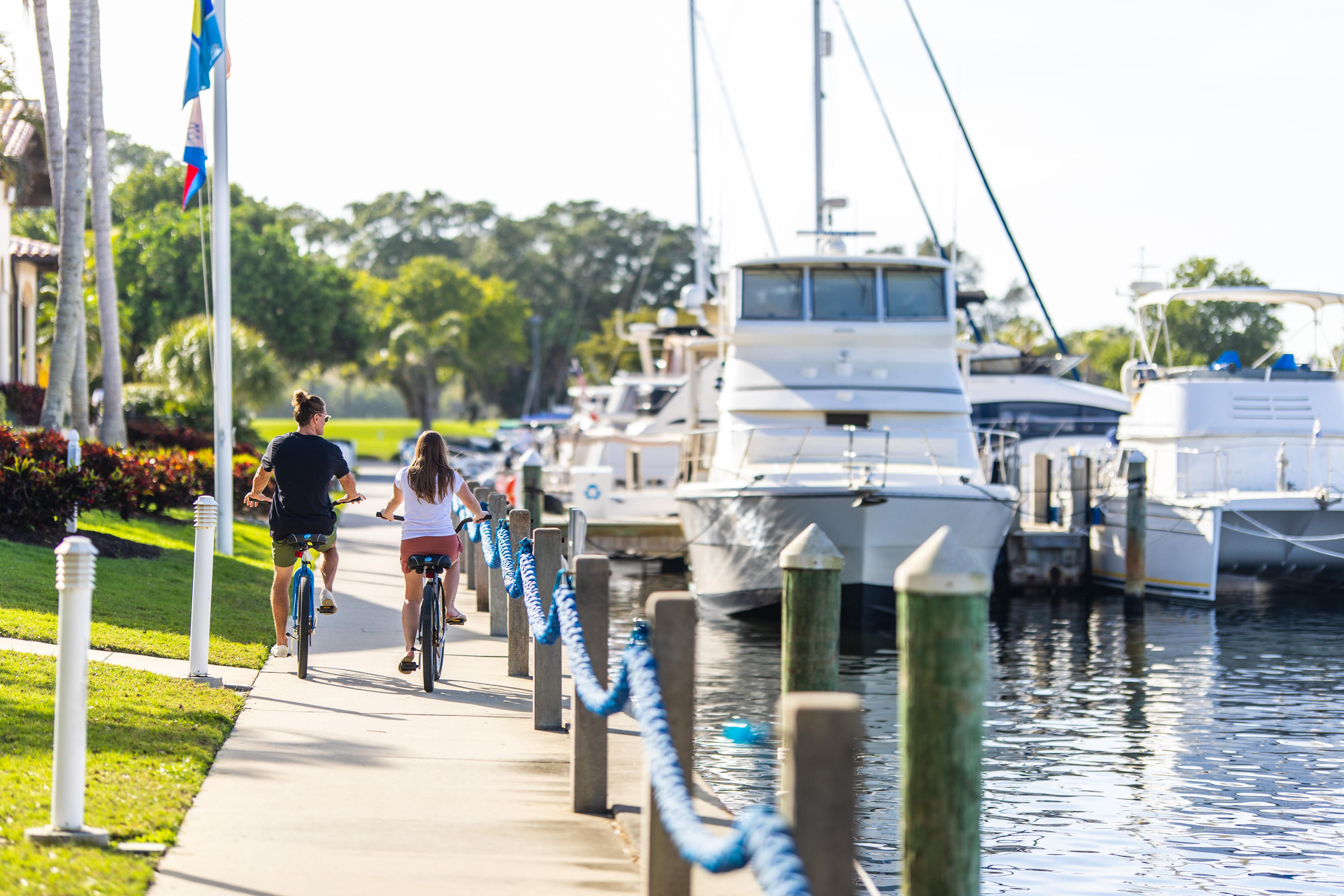 Couple riding bikes on a sidewalk next to large yachts docked at a marina