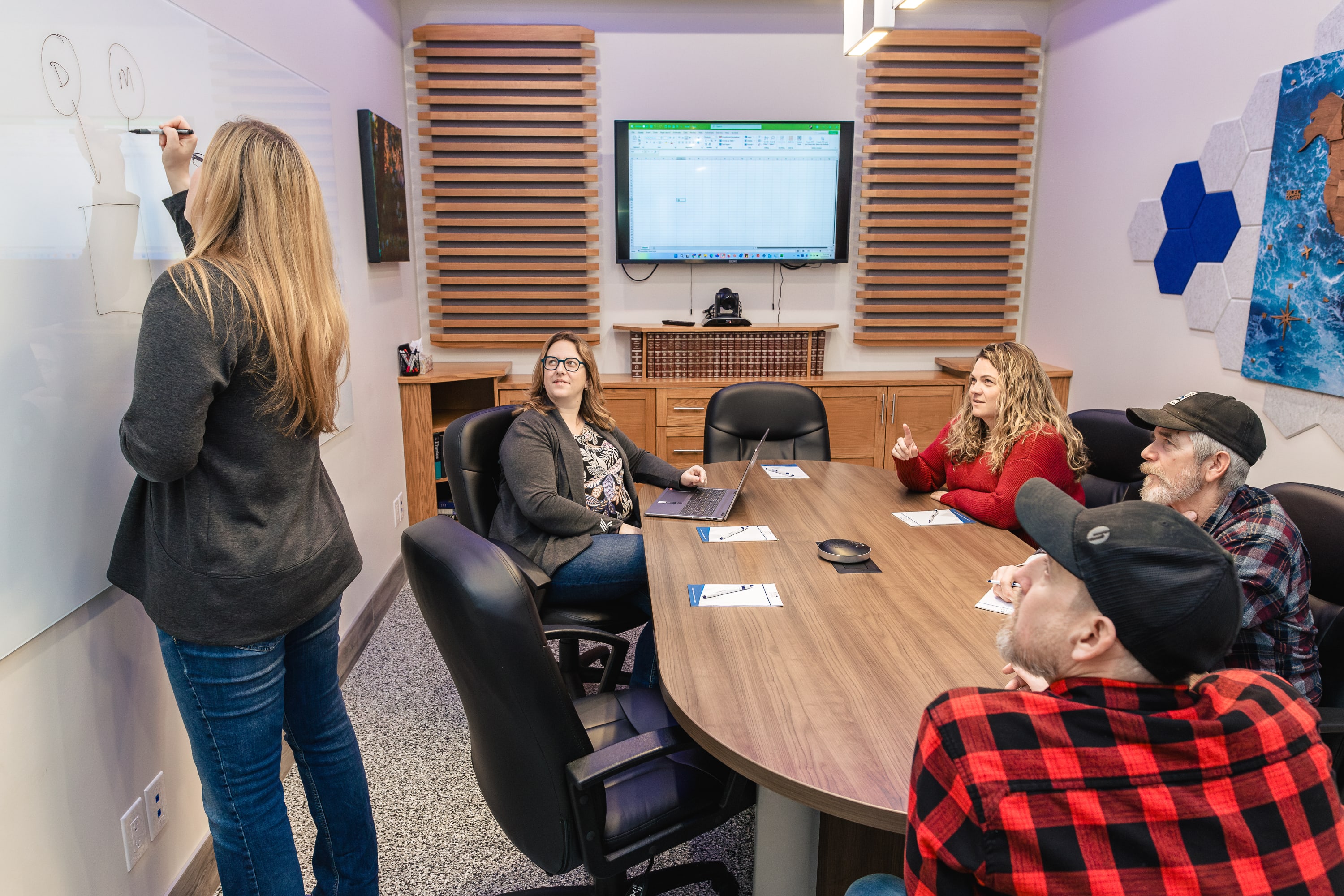 A woman stands at a whiteboard drawing a diagram, while four seated individuals around a conference table observe and engage.