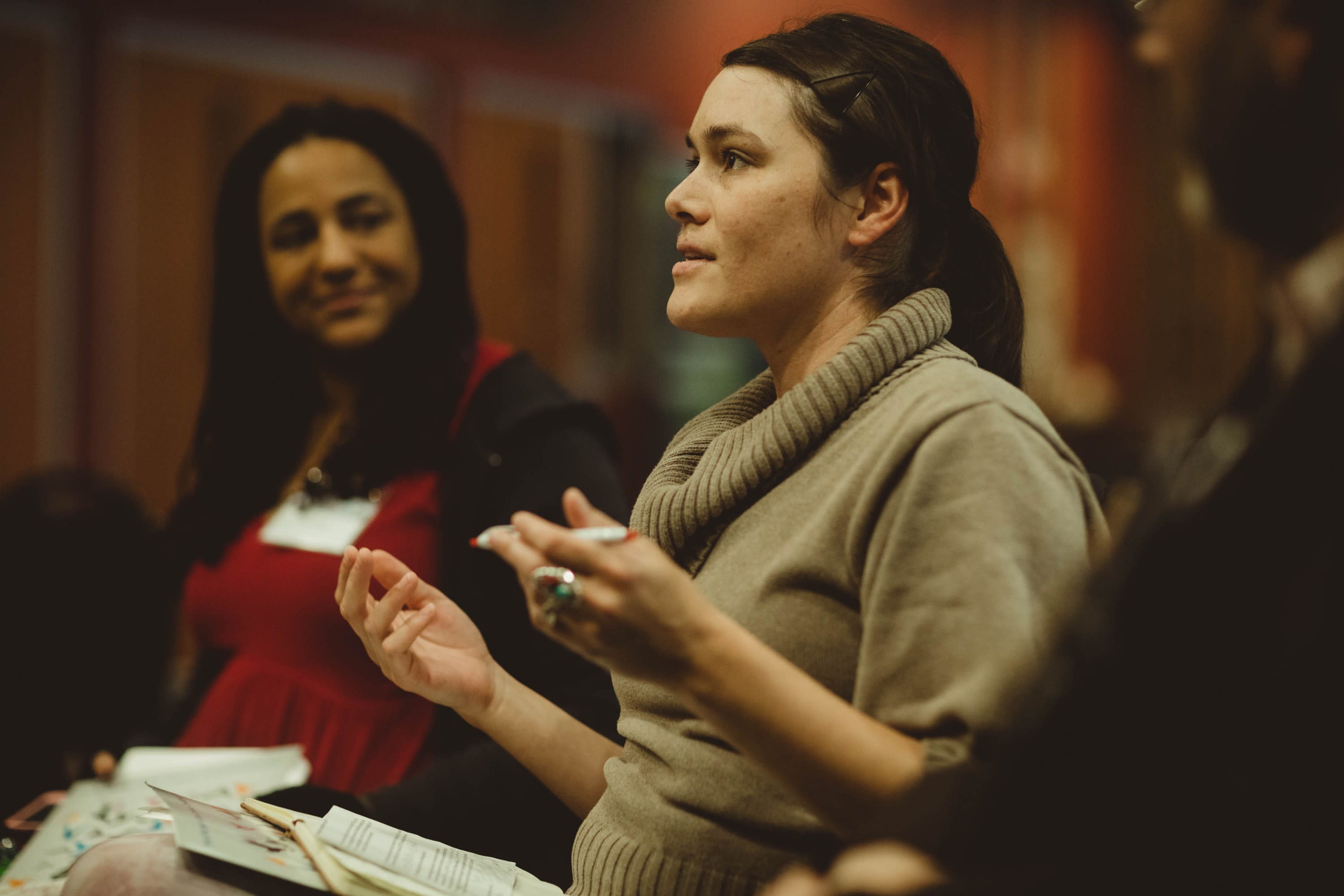 A speaker is seated, gesturing thoughtfully. Other speakers seated nearby listen intently.