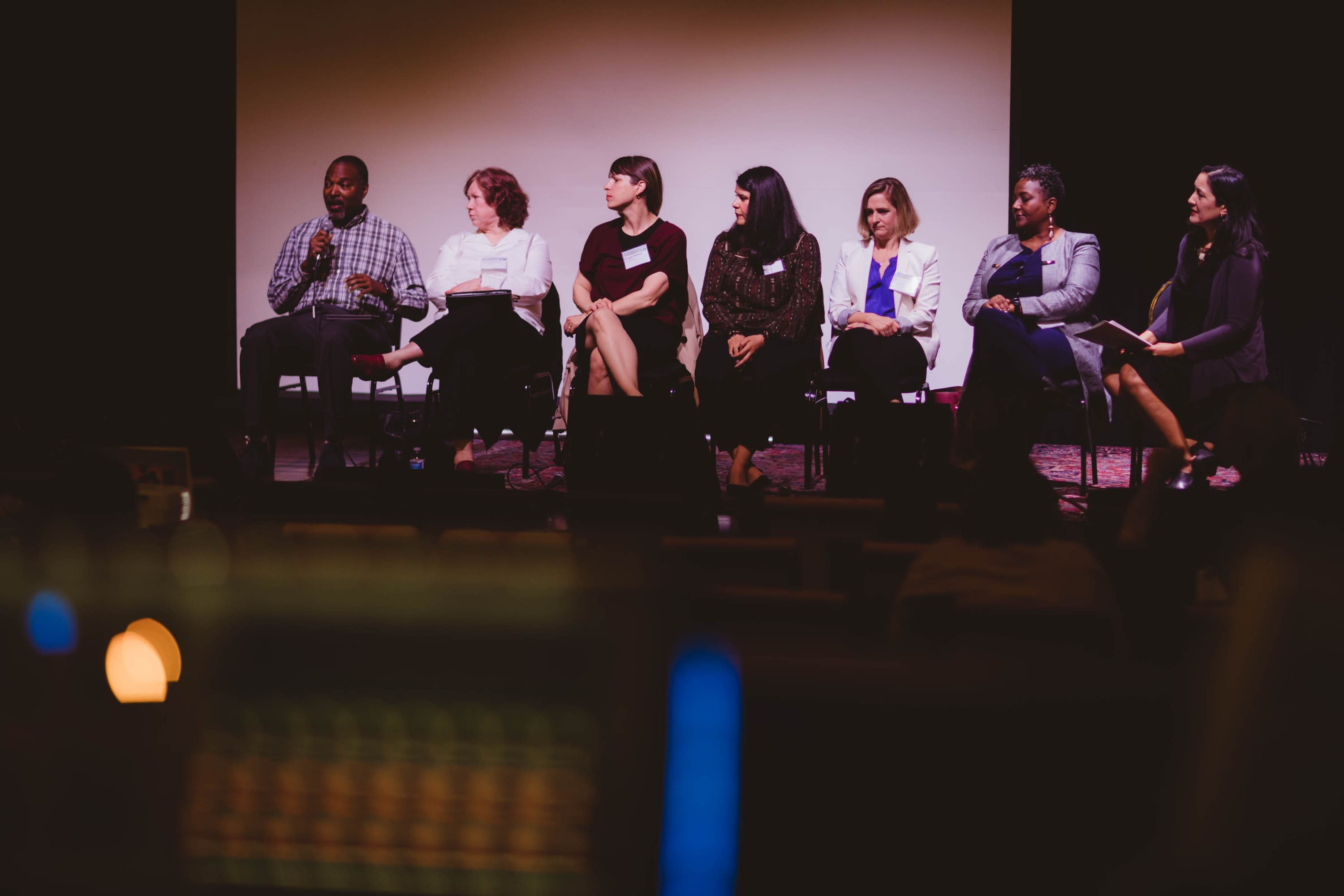 A diverse group of seven panelists are seated on the stage.