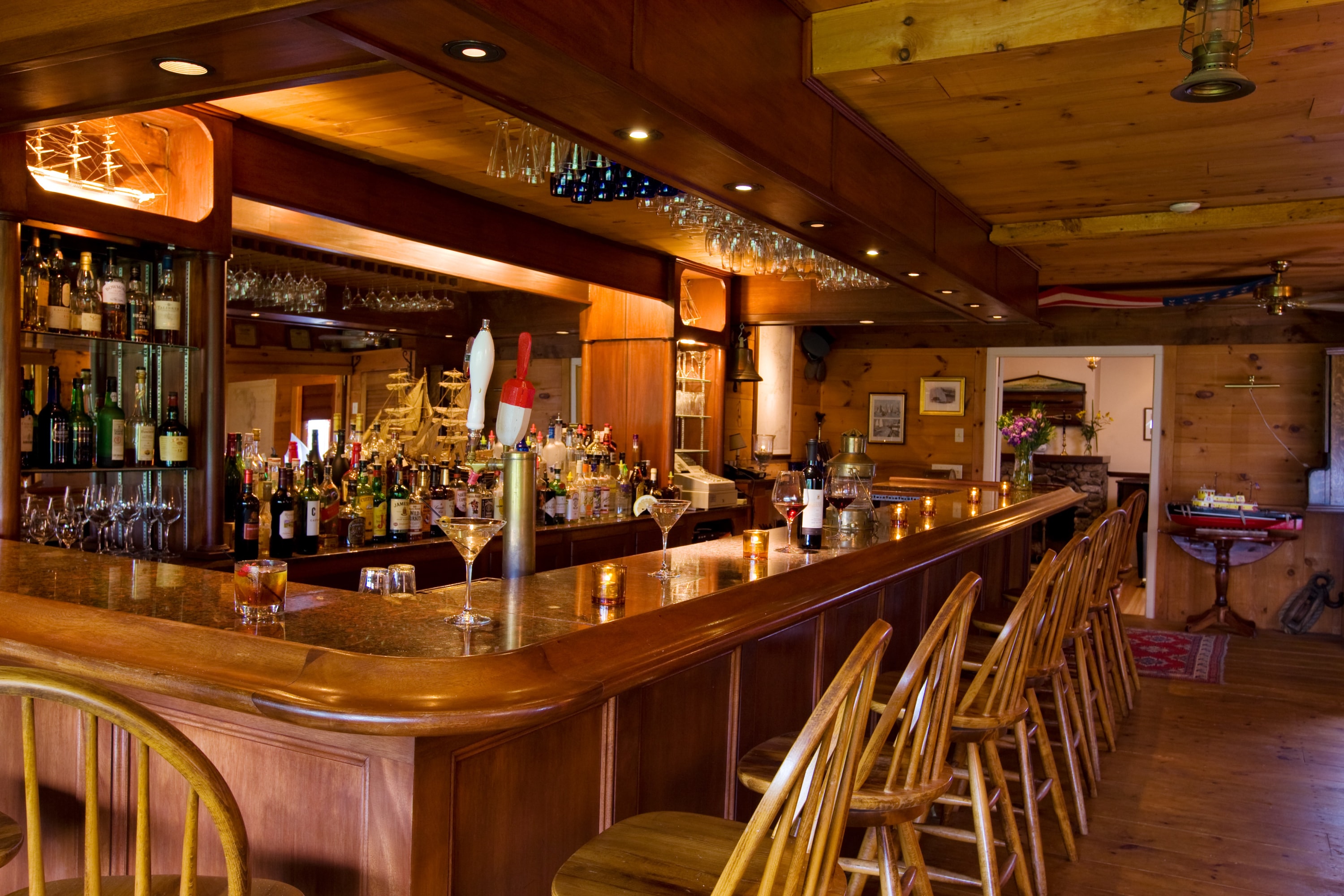 A warm bar, surrounded by wood with chairs bellied up to the bar, mirror reflecting behind the bar with liquor bottles.