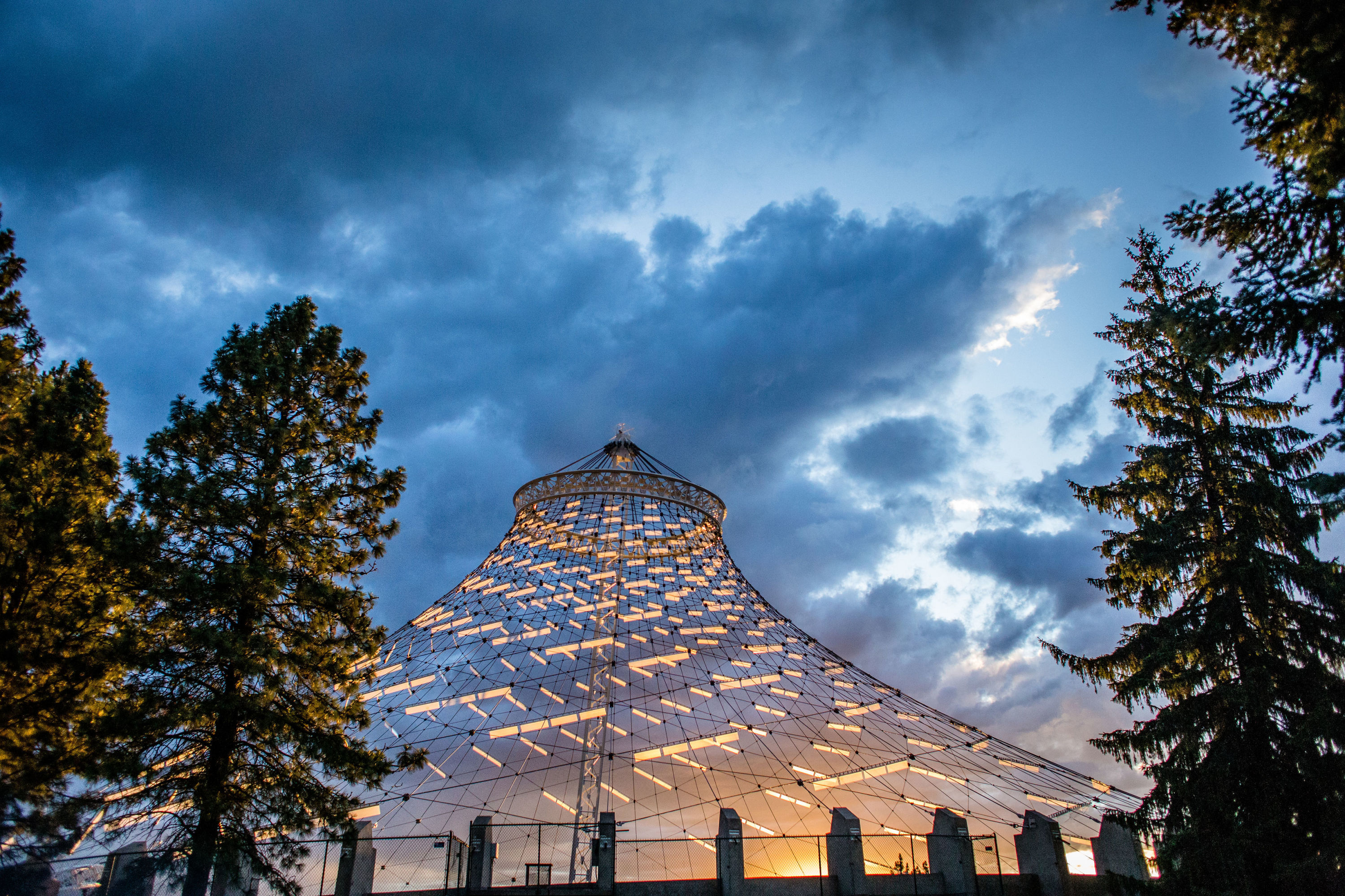 Sunset picture of the Pavilion structure looking up from the ground with trees flanking both sides and cloudy skies.