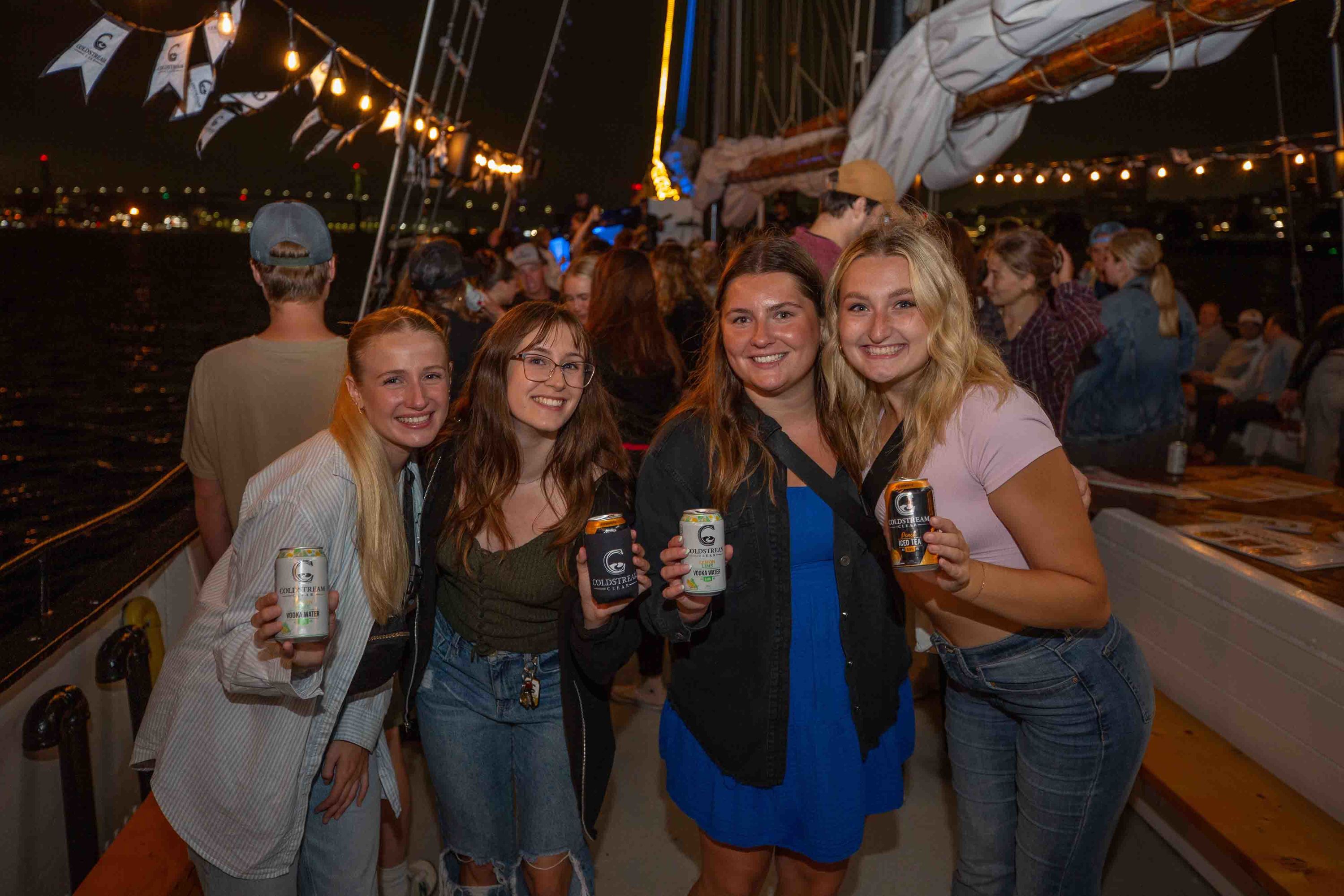 Four friends smile as they raise their cans of Coldstream beverages on the Coldstream Party Boat