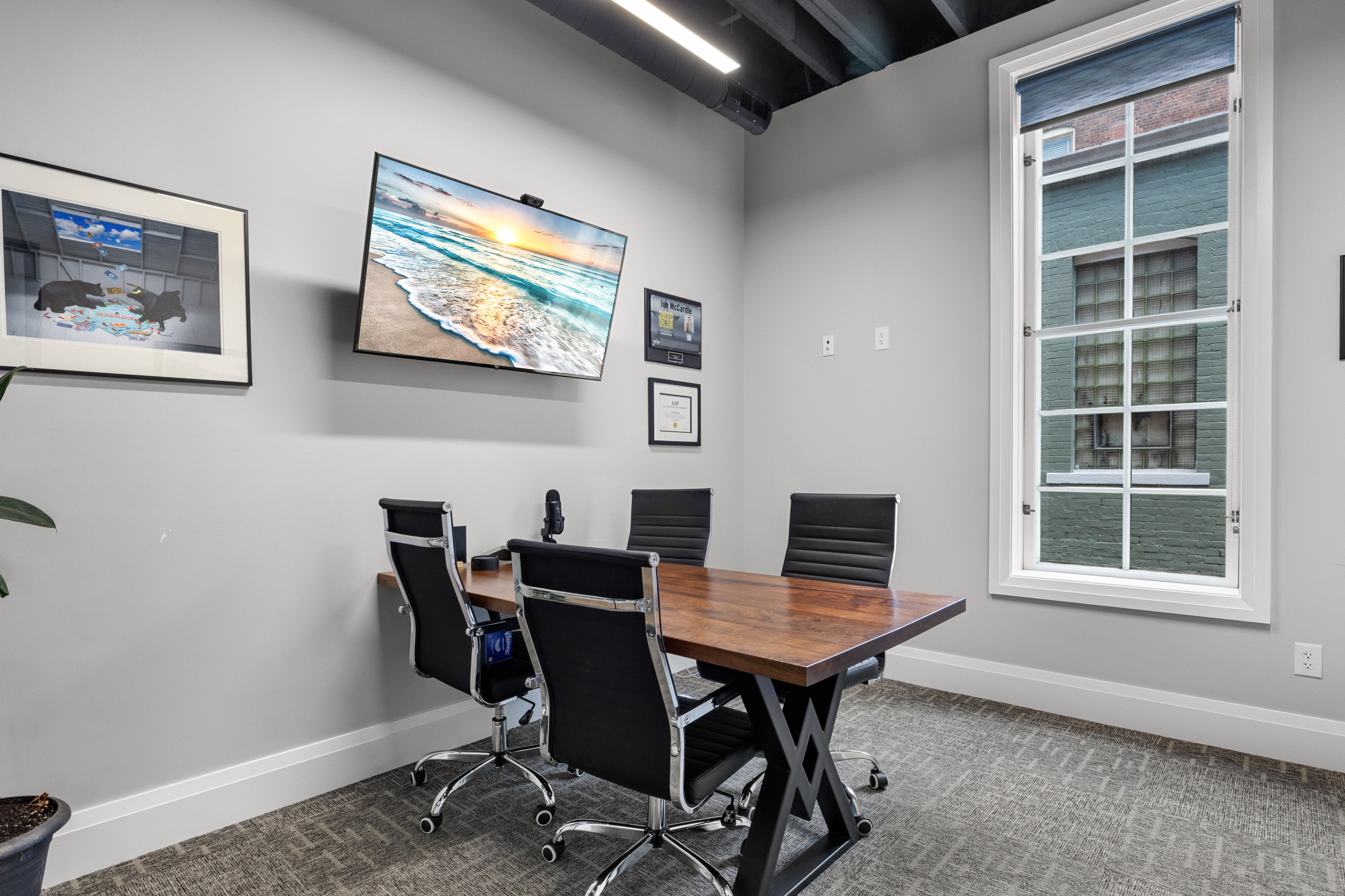 Conference room with a wooden table, four black chairs, a TV displaying a beach scene, framed art and certificates, and a gla