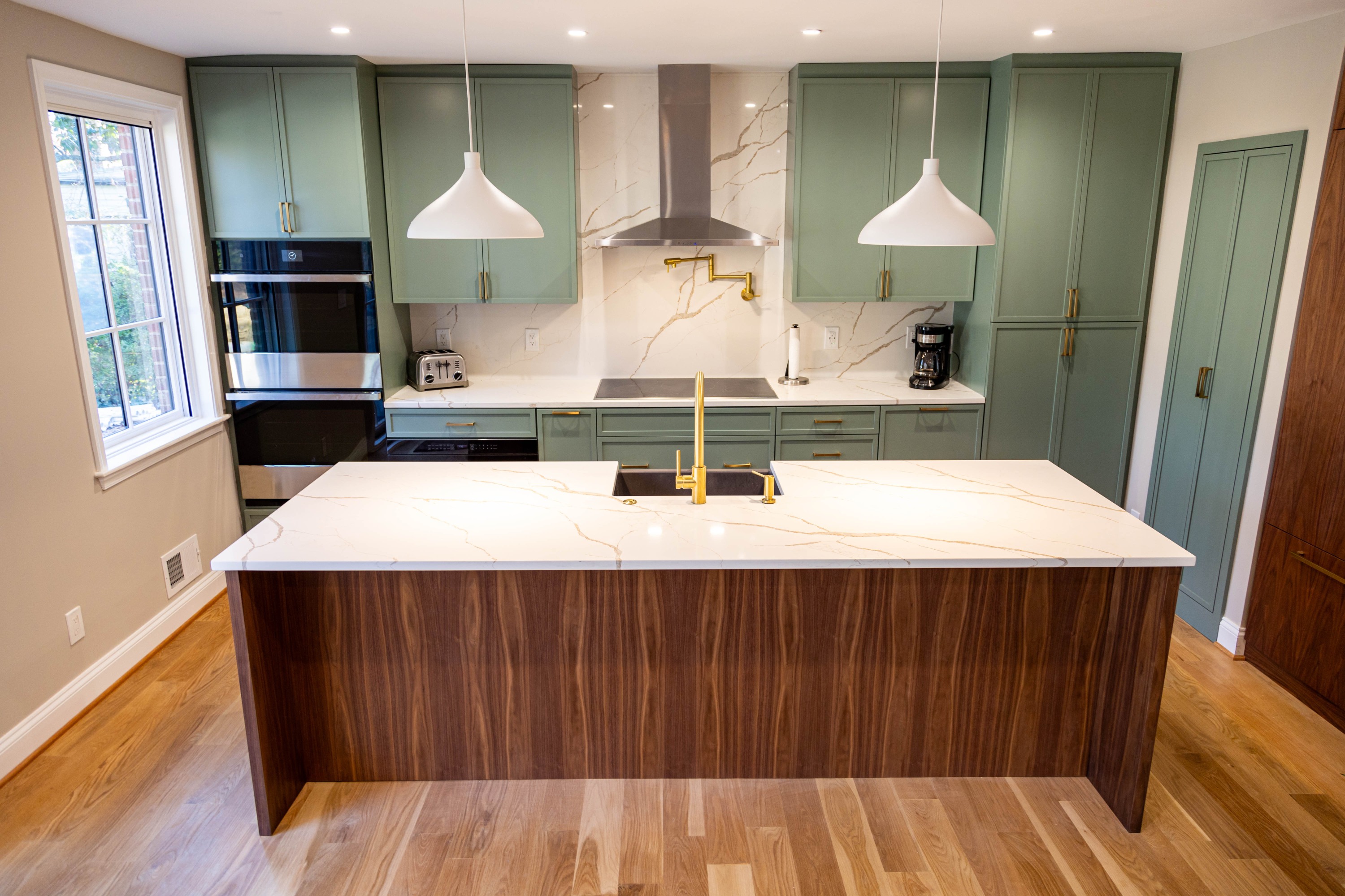 A modern kitchen featuring sage green upper and lower cabinets, a large central island with a waterfall edge in rich walnut
