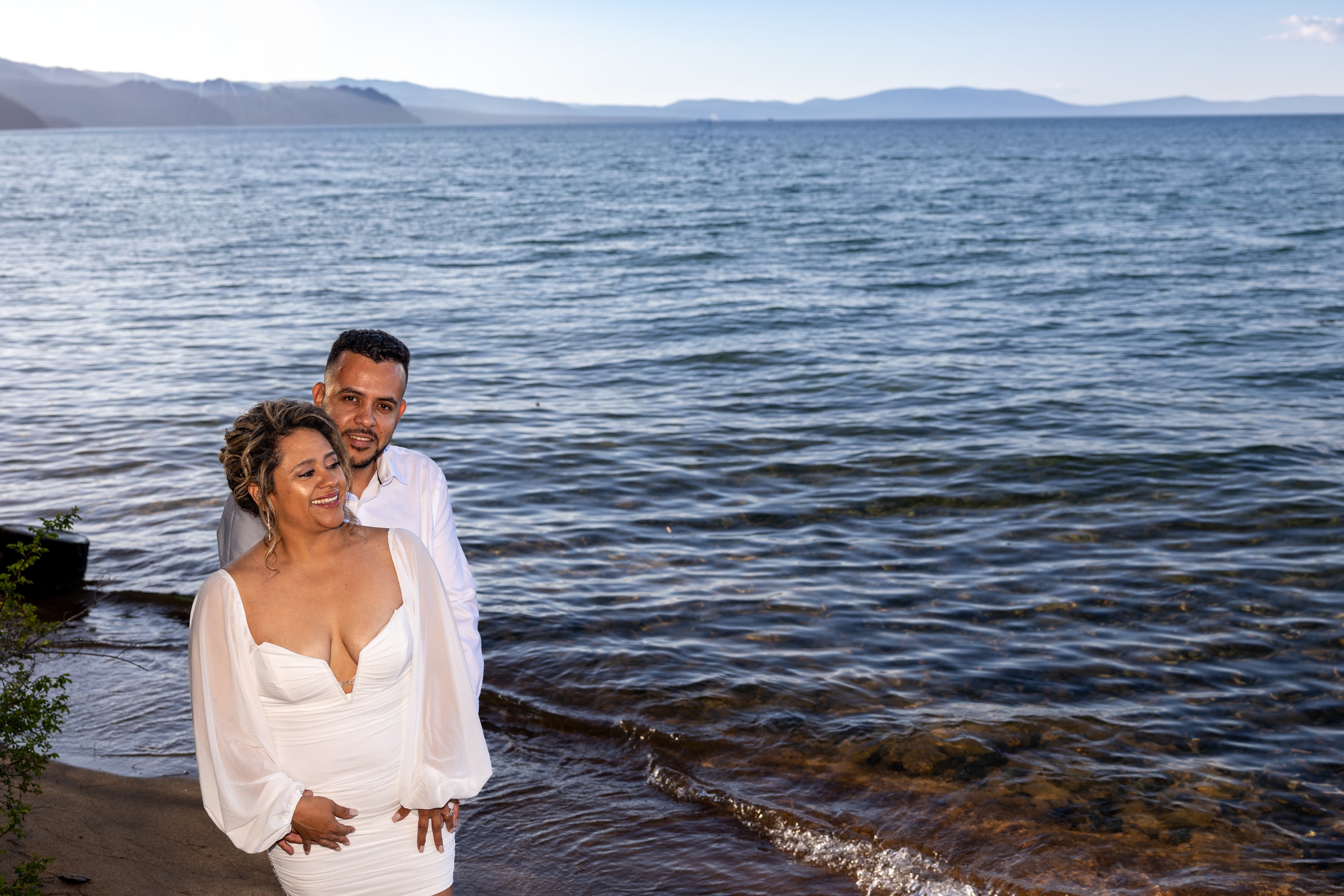 Smiling couple stands at the edge of Lake Tahoe, with the bride in a white dress and the groom in a white shirt, framed by so