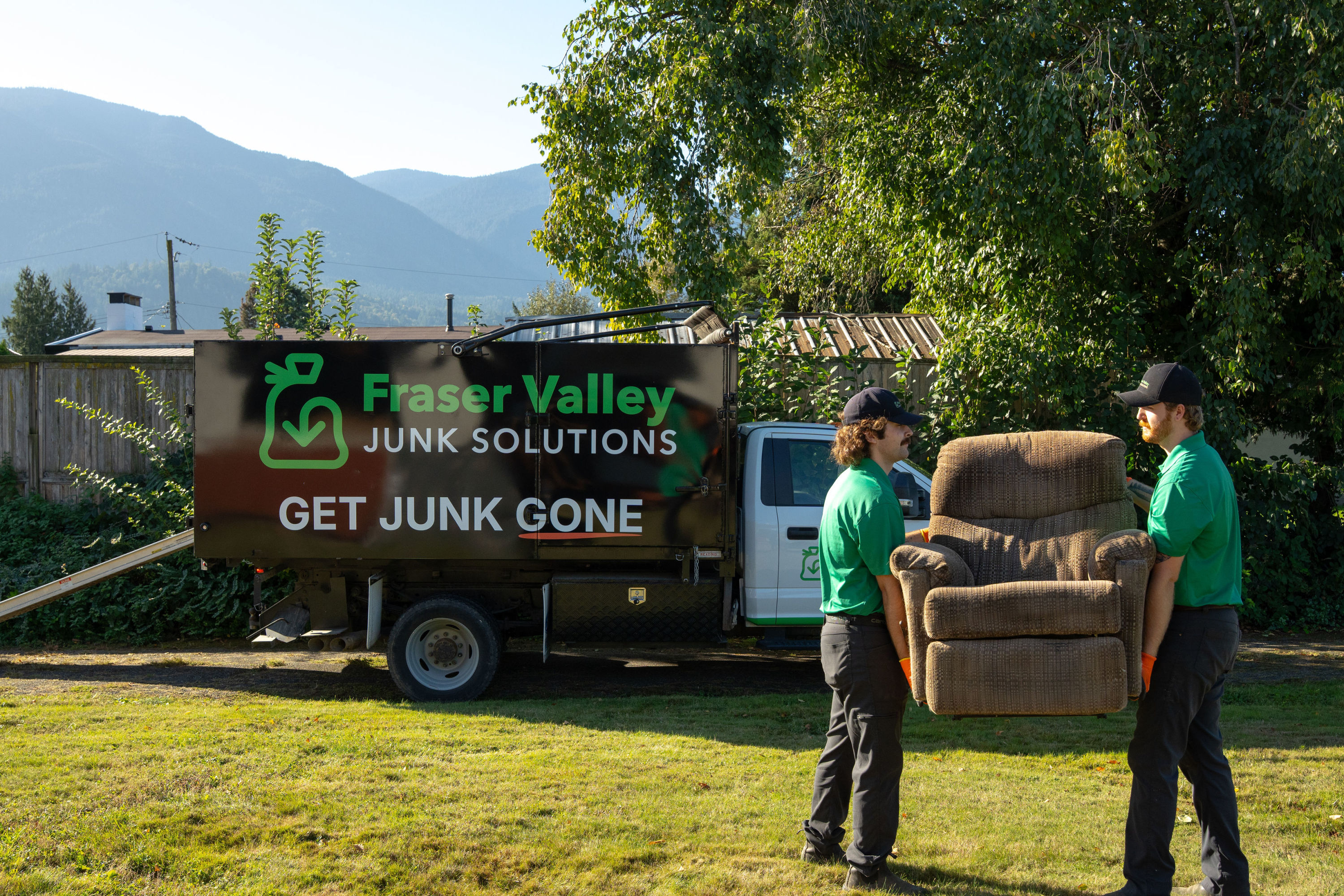 Two Fraser Valley Junk Solutions employees carrying a recliner to the FVJS truck for disposal in Chilliwack, with clear skies