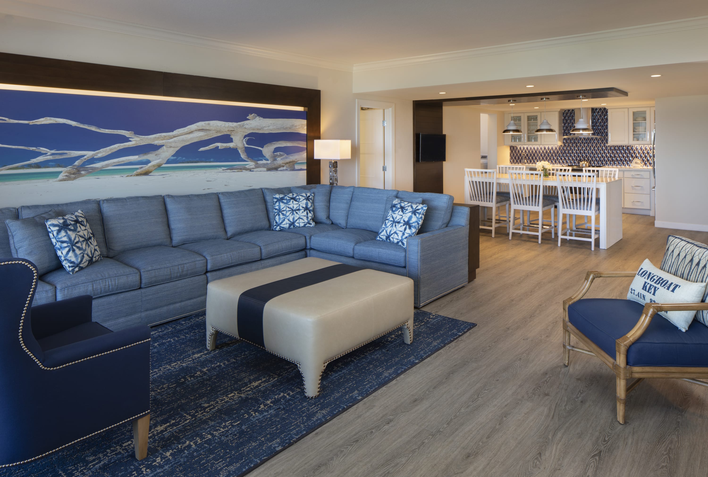 Blue "L" shaped couch with oversized ottoman, wingback chair, armchair with pillow and dining table and kitchen in background