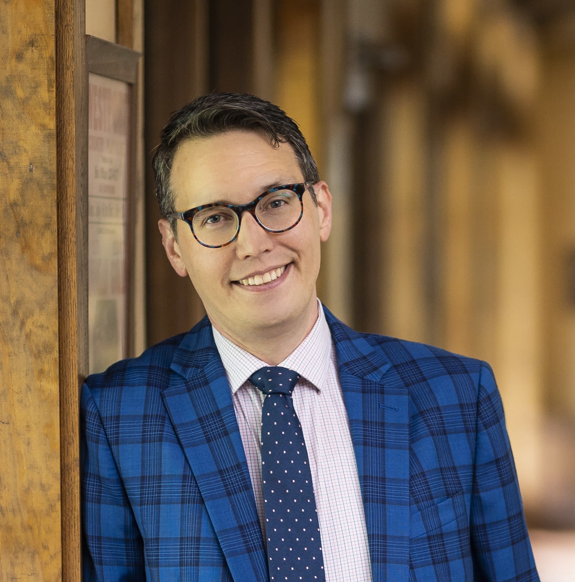 A photo of a man wearing glasses in a plaid blue suit with a blue tie leaning on a wooden wall. He is Michael Barker.