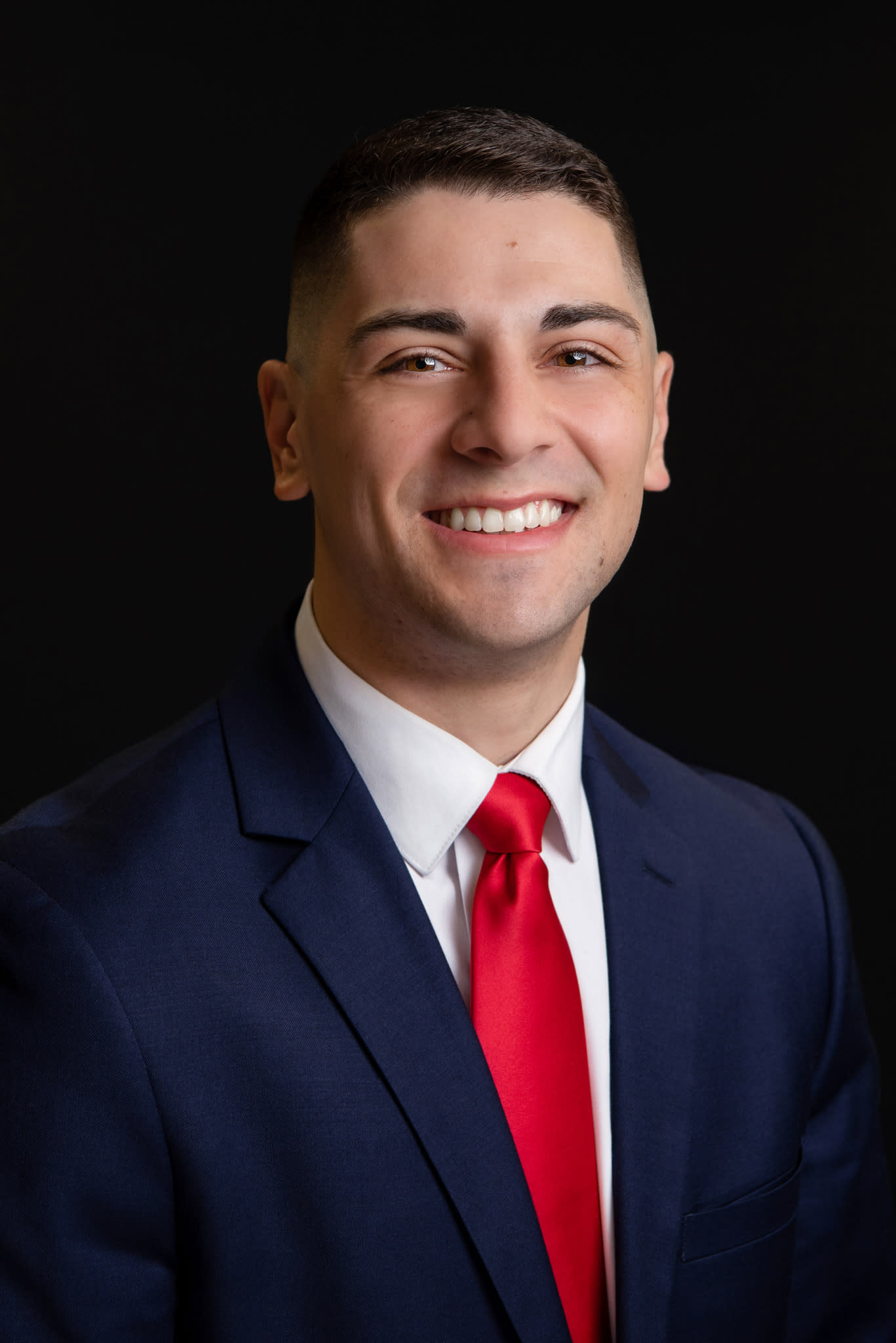 A Caucasian man wearing a navy blue suit, white shirt and red tie at his headshot session.