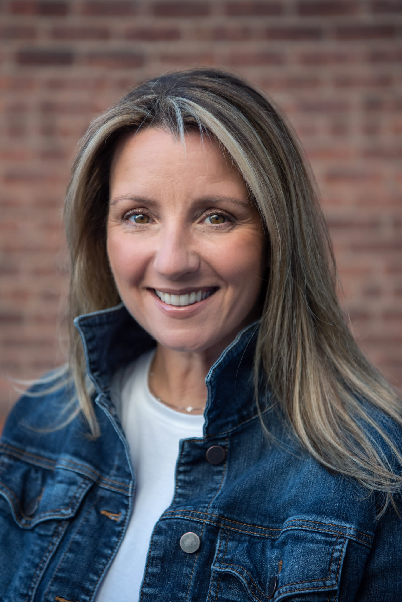 A Caucasian woman standing in front of a brick wall with a jean jacket and white shirt smiling for her headshot session.