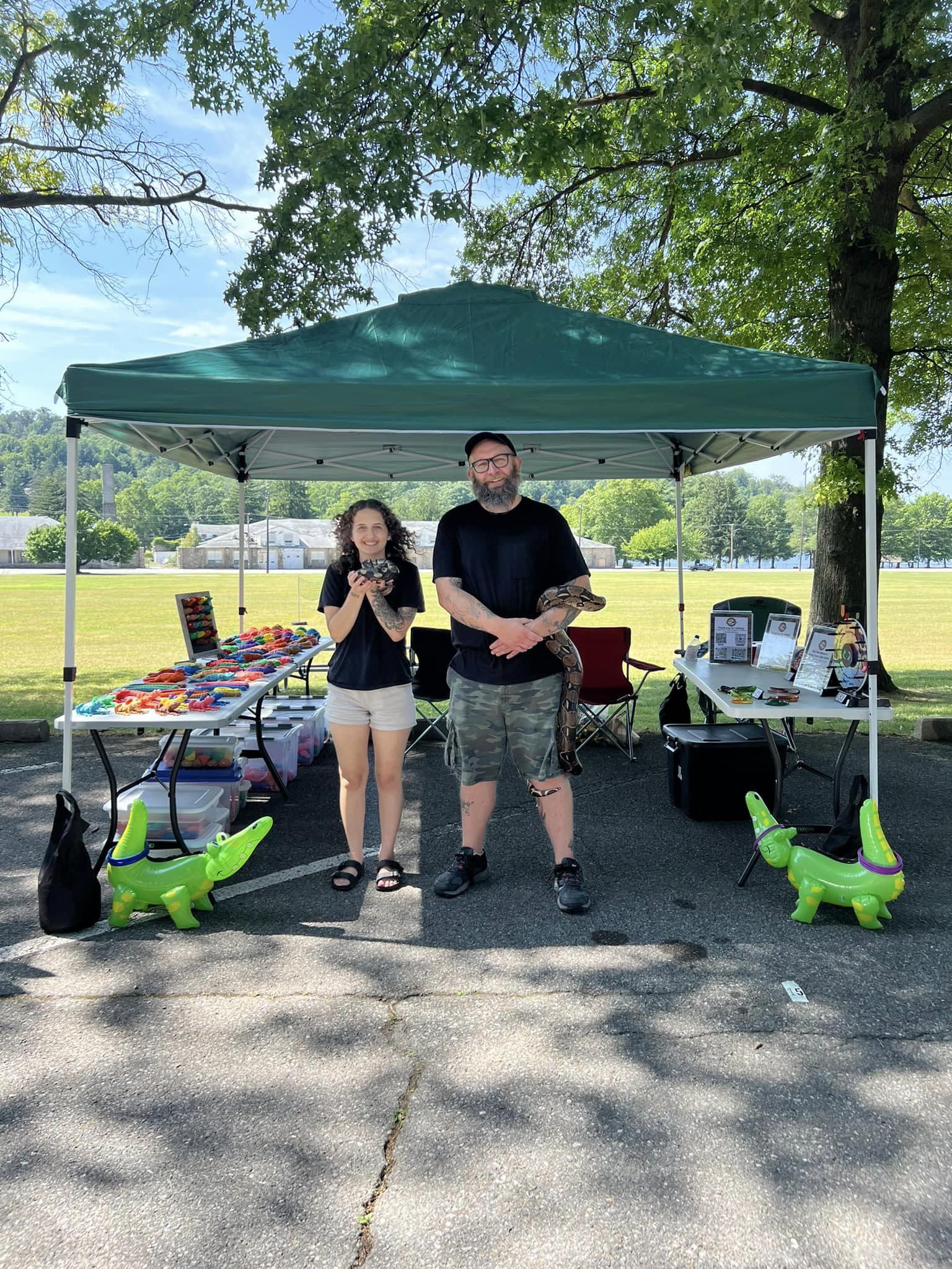 Two people standing under a green canopy holding reptiles, with tables displaying toys and materials, set in a sunny park