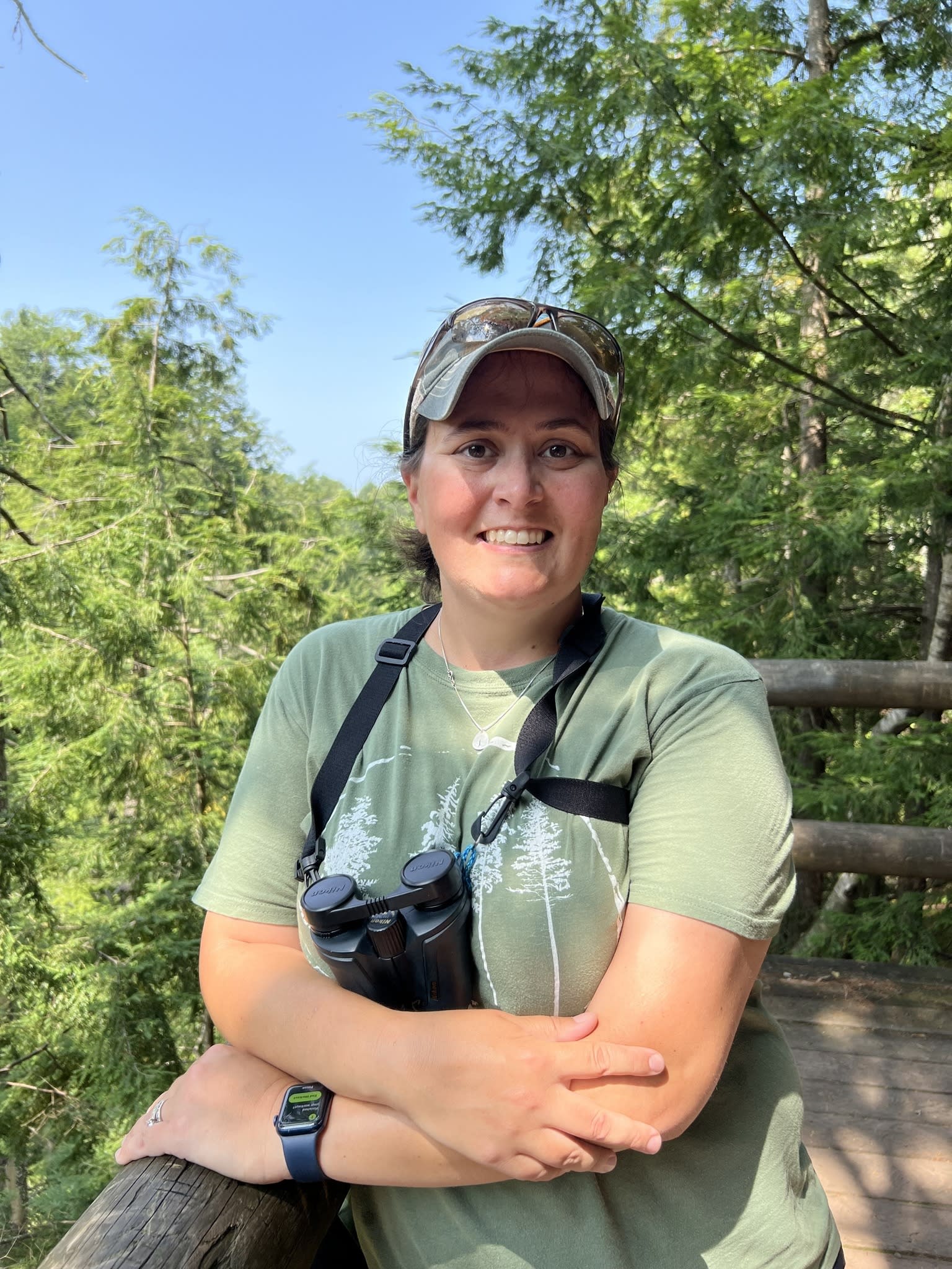 Sam standing on a wooden platform, in the woods, wearing a green shirt, binoculars and a baseball cap.