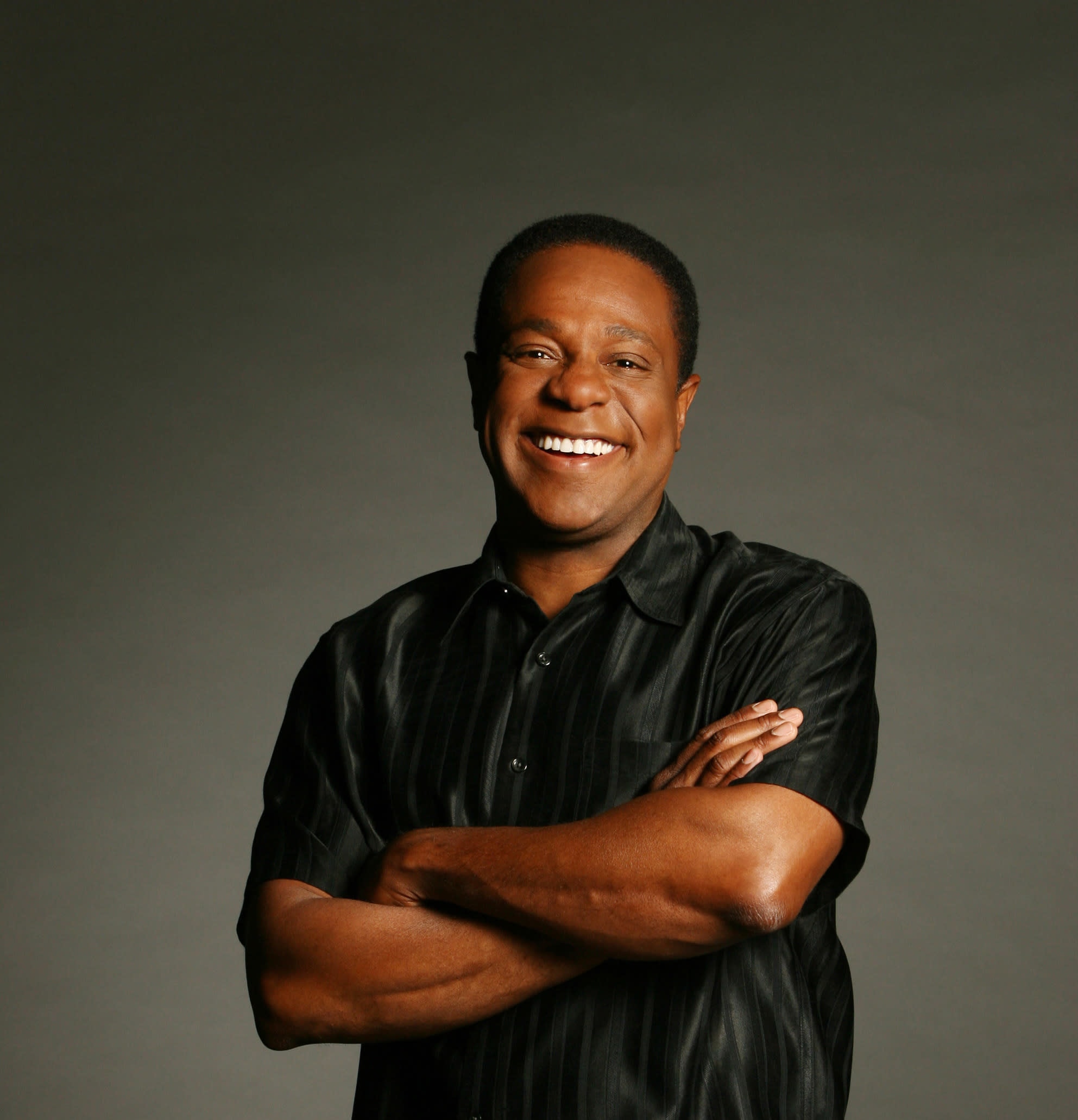 Smiling black man with short cropped hair with his arms crossed wearing a black satin polo shirt