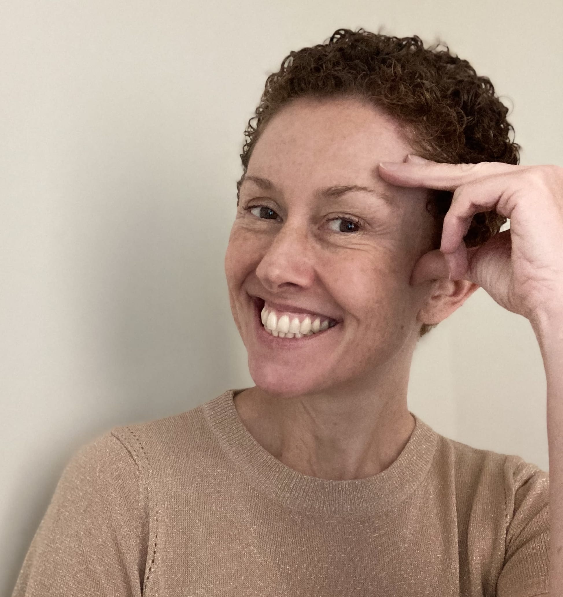 White women with short, curly auburn hair, smiling broadly and wearing a sparkly peach-toned sweater.