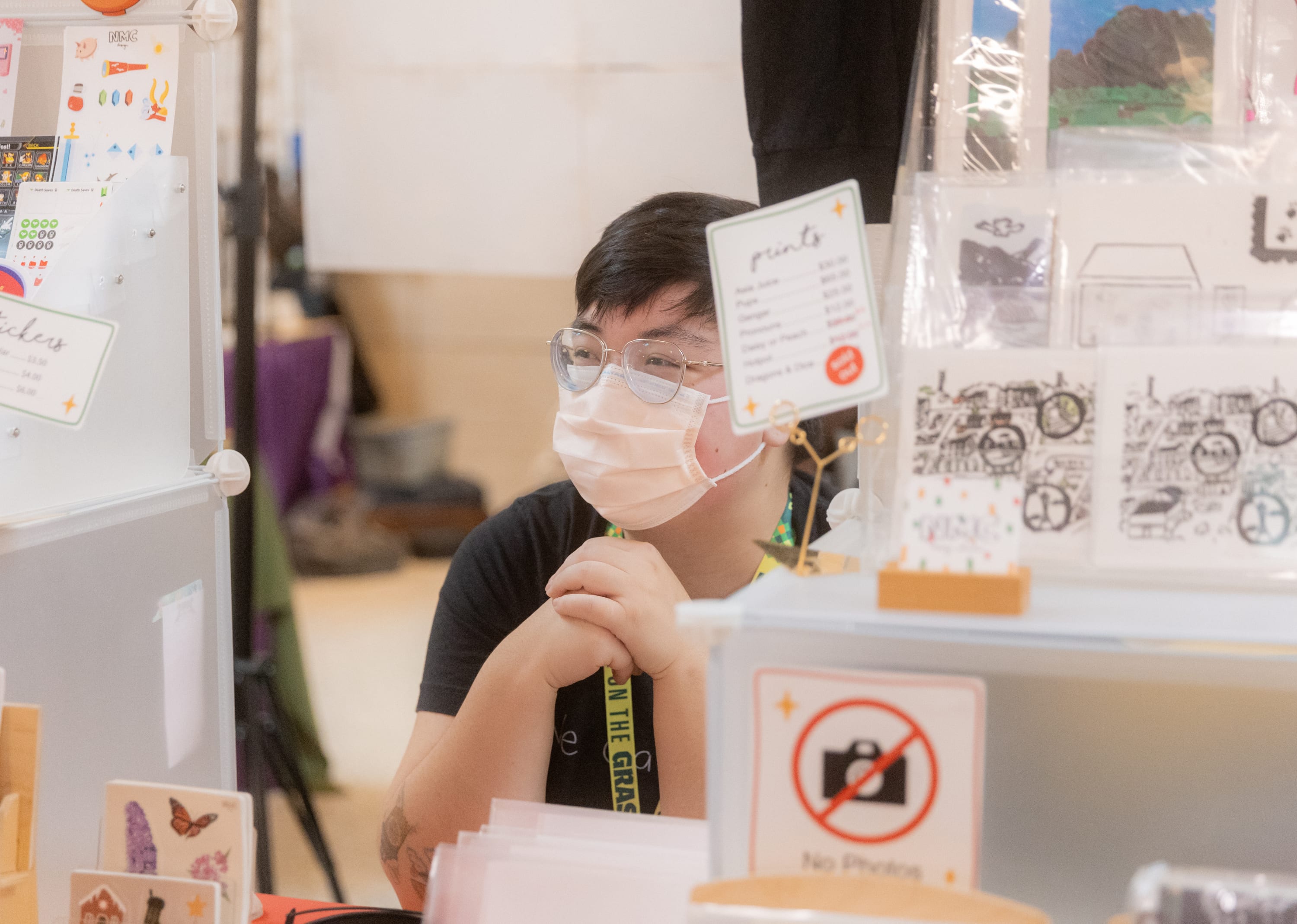Natalie, the artist is wearing a mask and sitting behind their booth table. There's some signage and prints on the table.