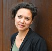 Portrait of woman with brown, short cropped hair with black jacket leaning against red iron wall, looks at camera