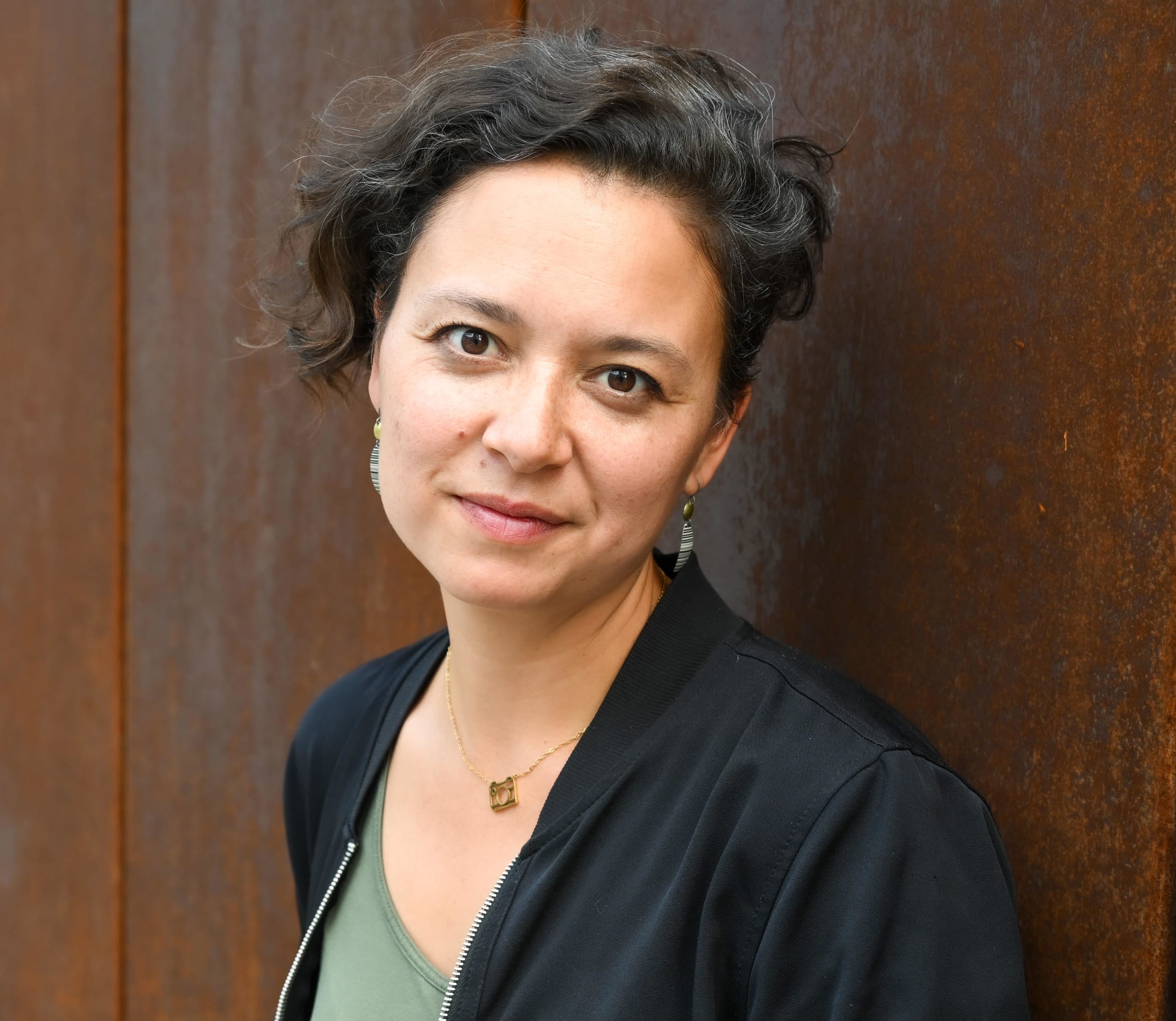 woman with short cut brown hair and brown eyes, looks at the camera with partial smile, leaning against red iron colored wall