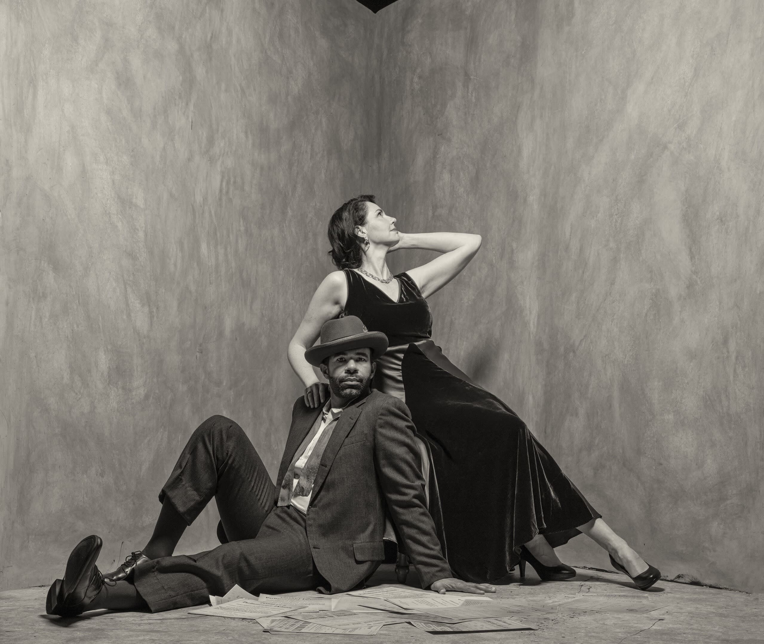 A black man in a suit sits on the floor leaning back against a stool. A white woman in a long dress splays across the stool.