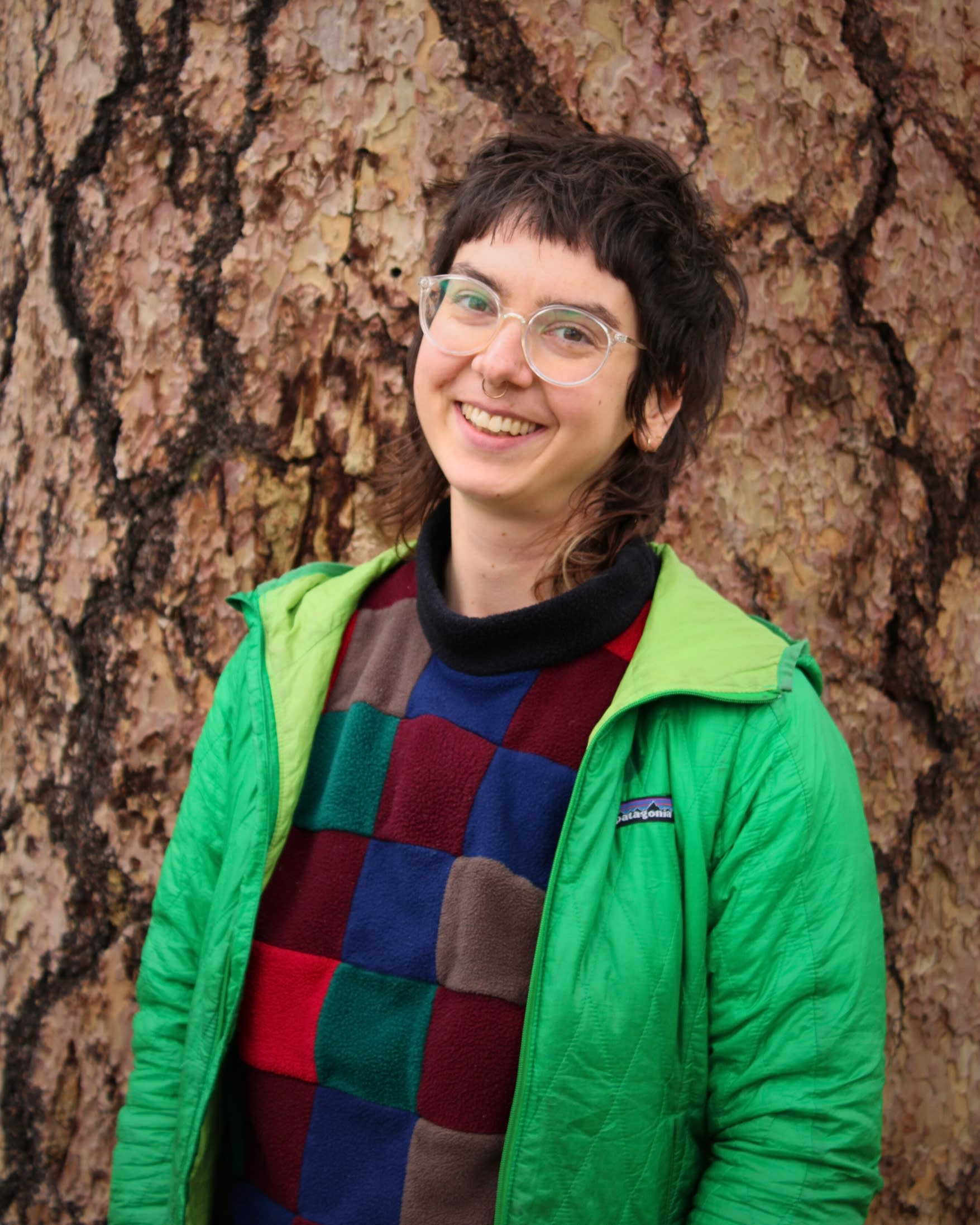 image of leah smiling, wearing a green jacket, leaning against a tree. they have clear glasses, a mullet, and a septum ring