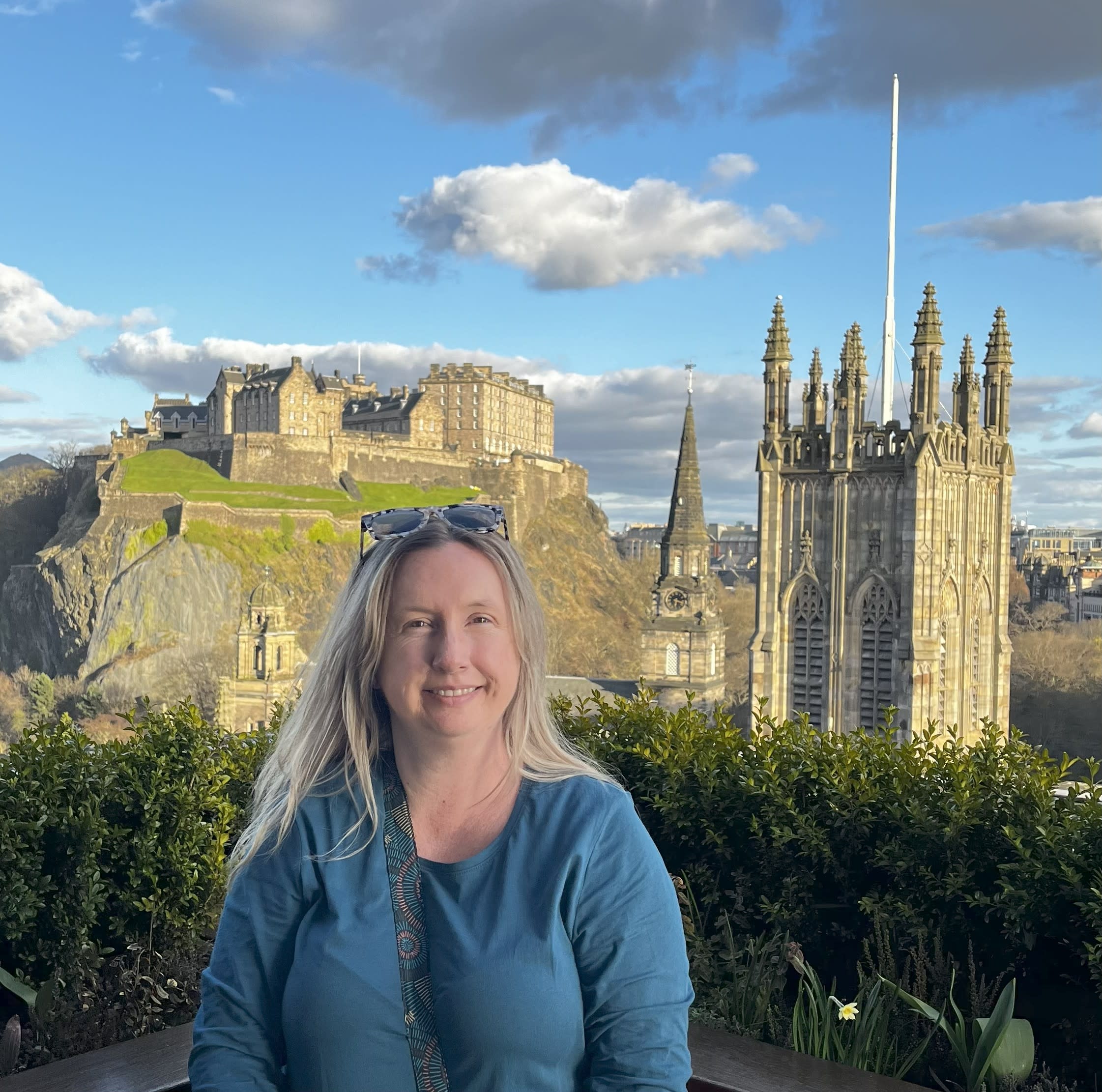 Stephanie smiling in front of Edinburgh Castle and historic church spires, with blue skies and clouds overhead.
