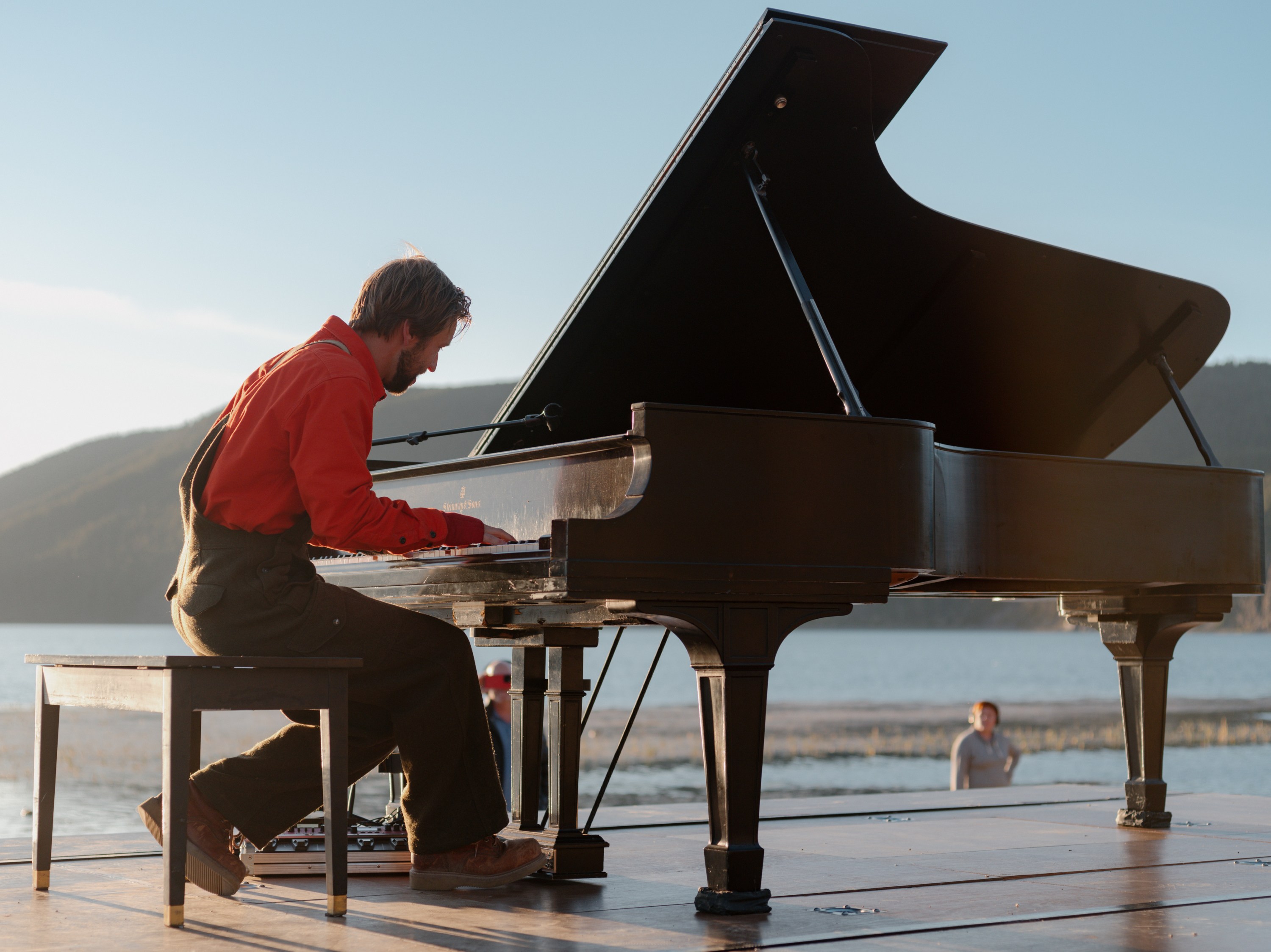 Hunter Noack plays piano infront of a lake a man stands in the background with headphones on listening to Hunter play