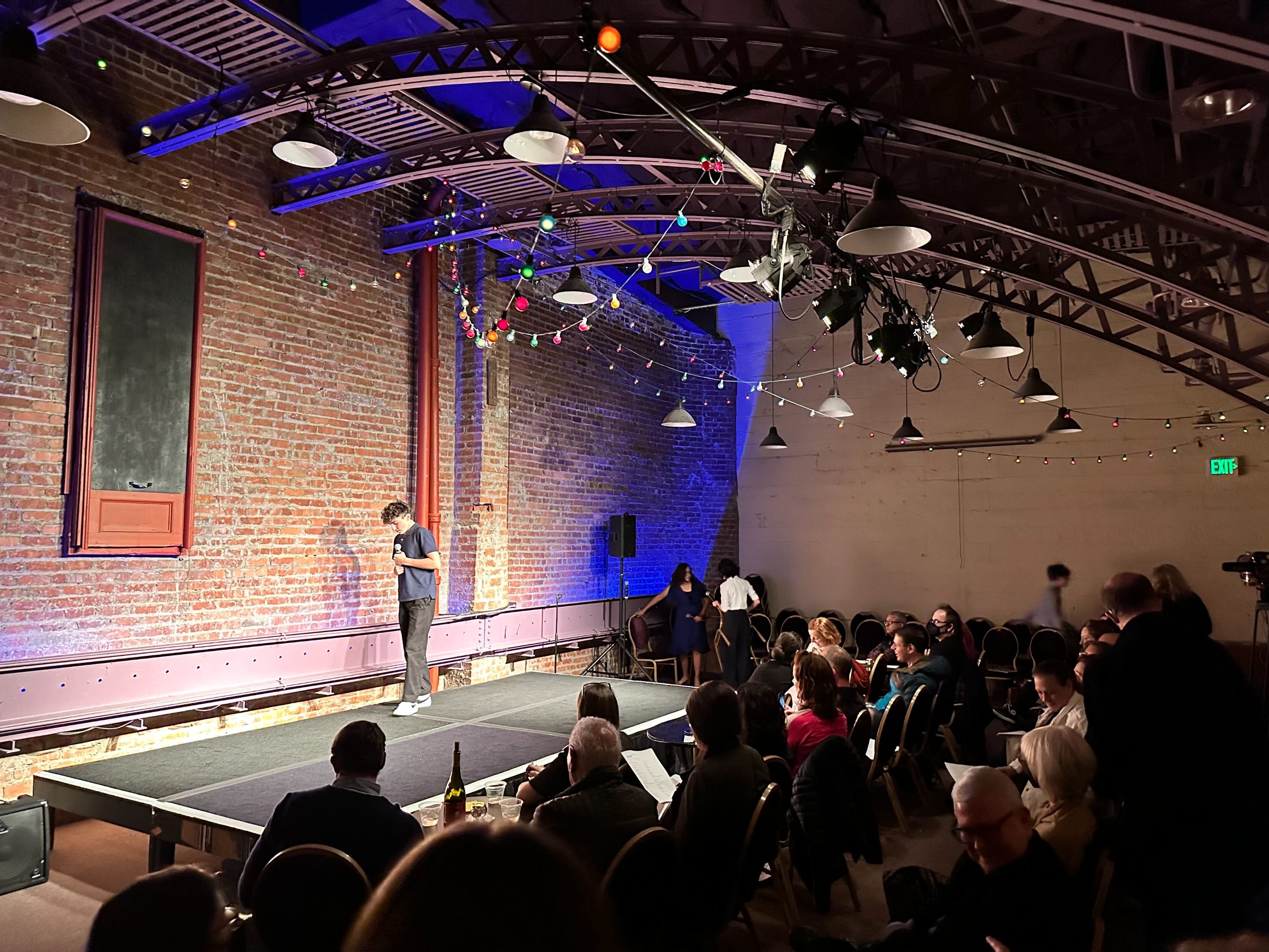 A figure speaking on stage in front of an intimate audience. The building has an industrial feel.