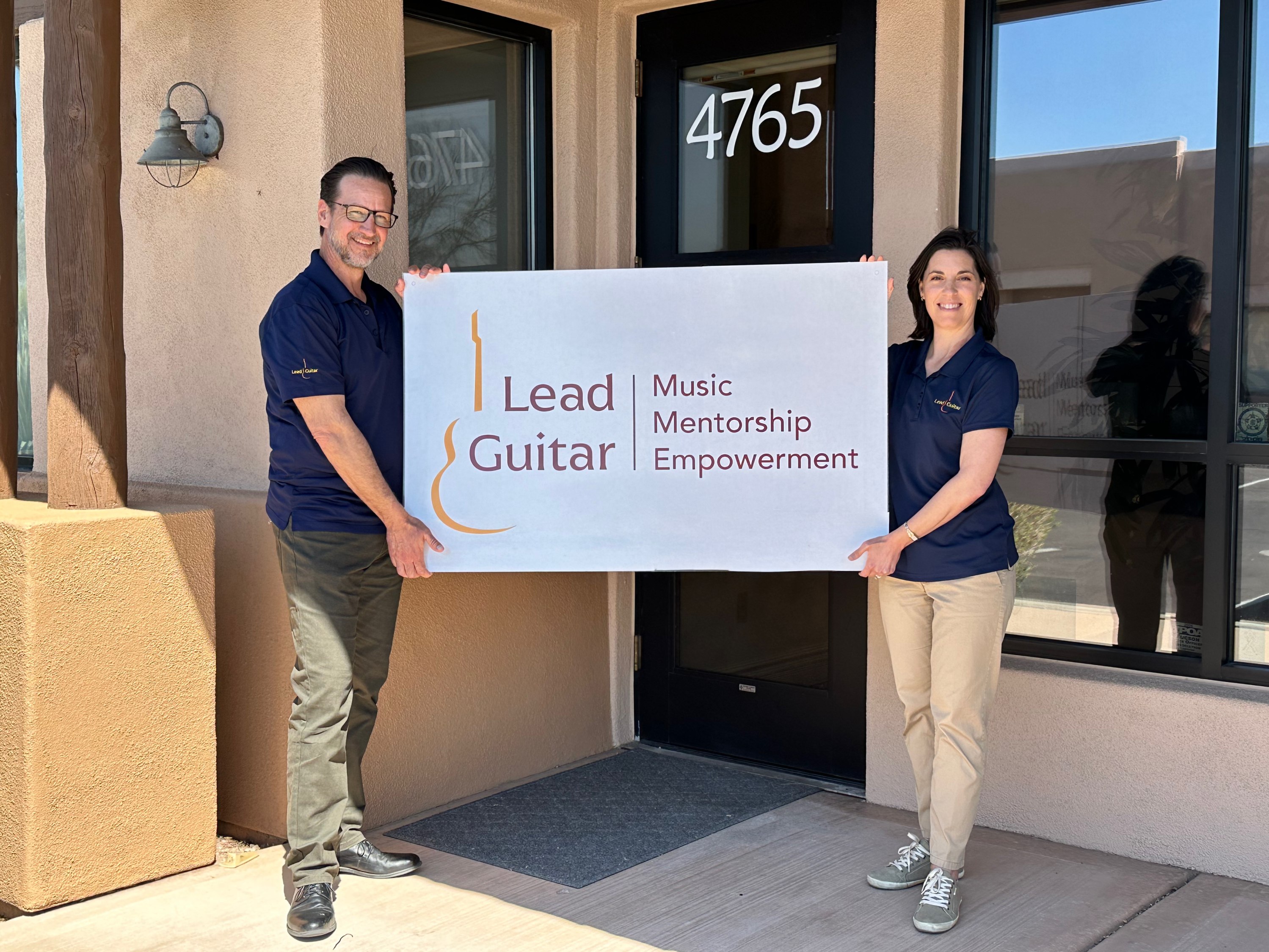 Lead Guitar staff outside the front door of their new building holding a sign