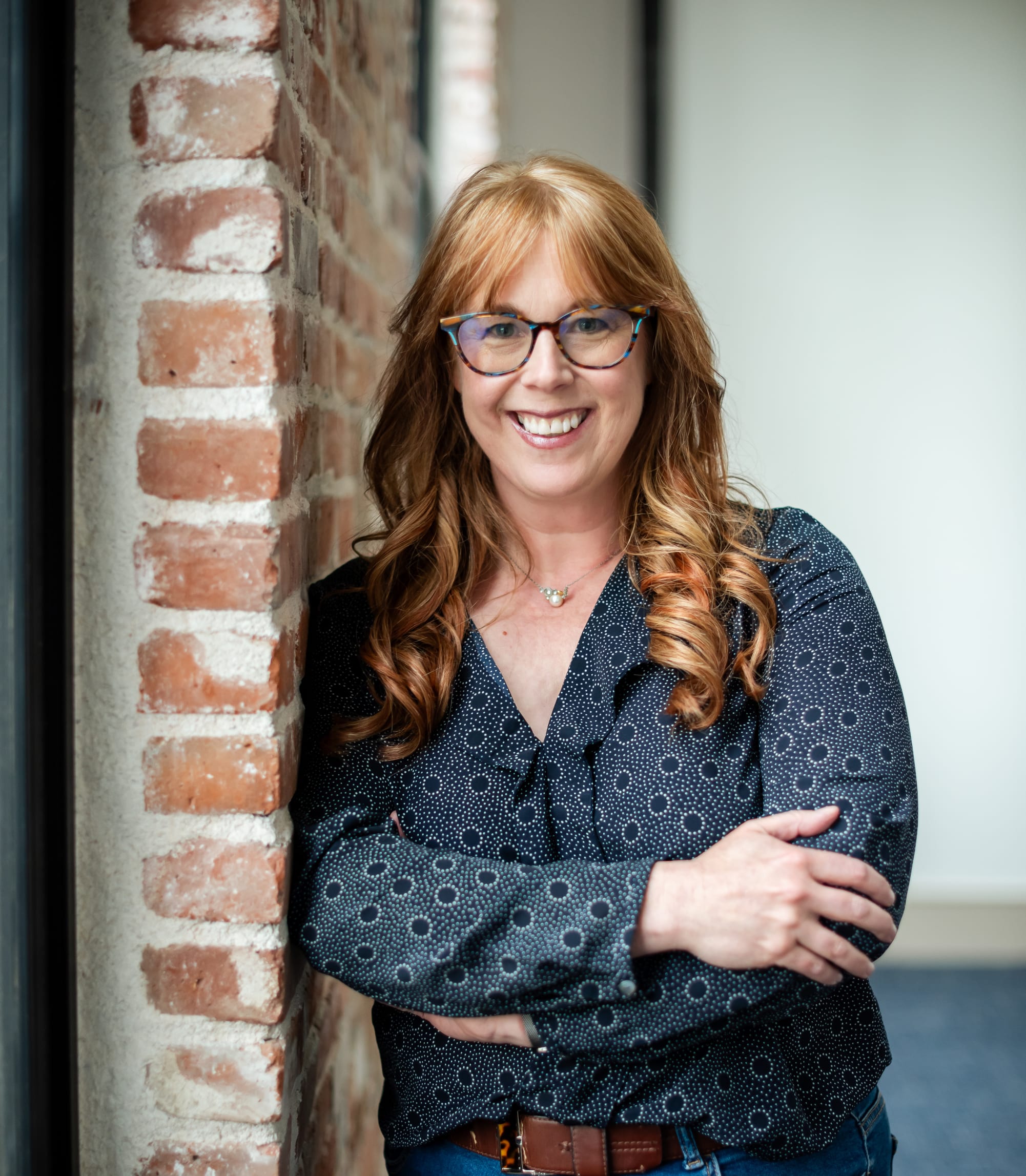 Catherine a woman with glasses and long reddish hair smiles and leans against a brick wall in a historic Longmont building