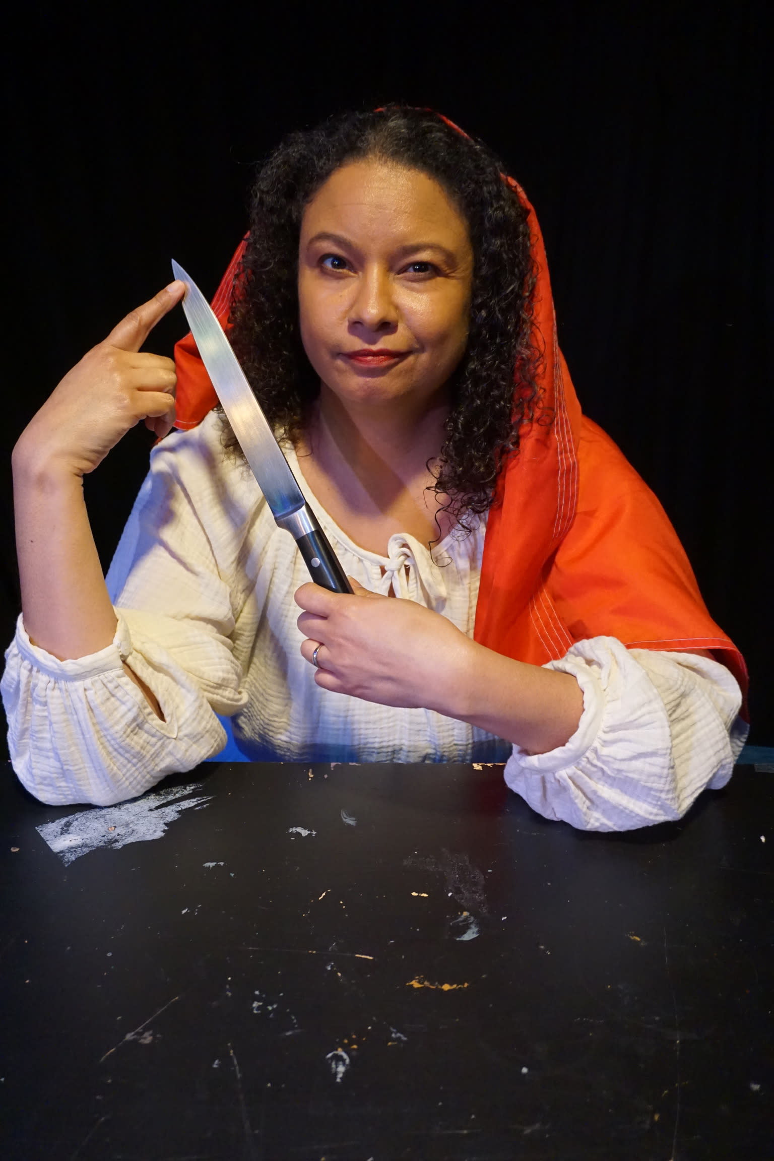 A black woman (Jennifer Greene) with her hair down, draped in a red scarf, wearing a white blouse and holding up a knife, loo