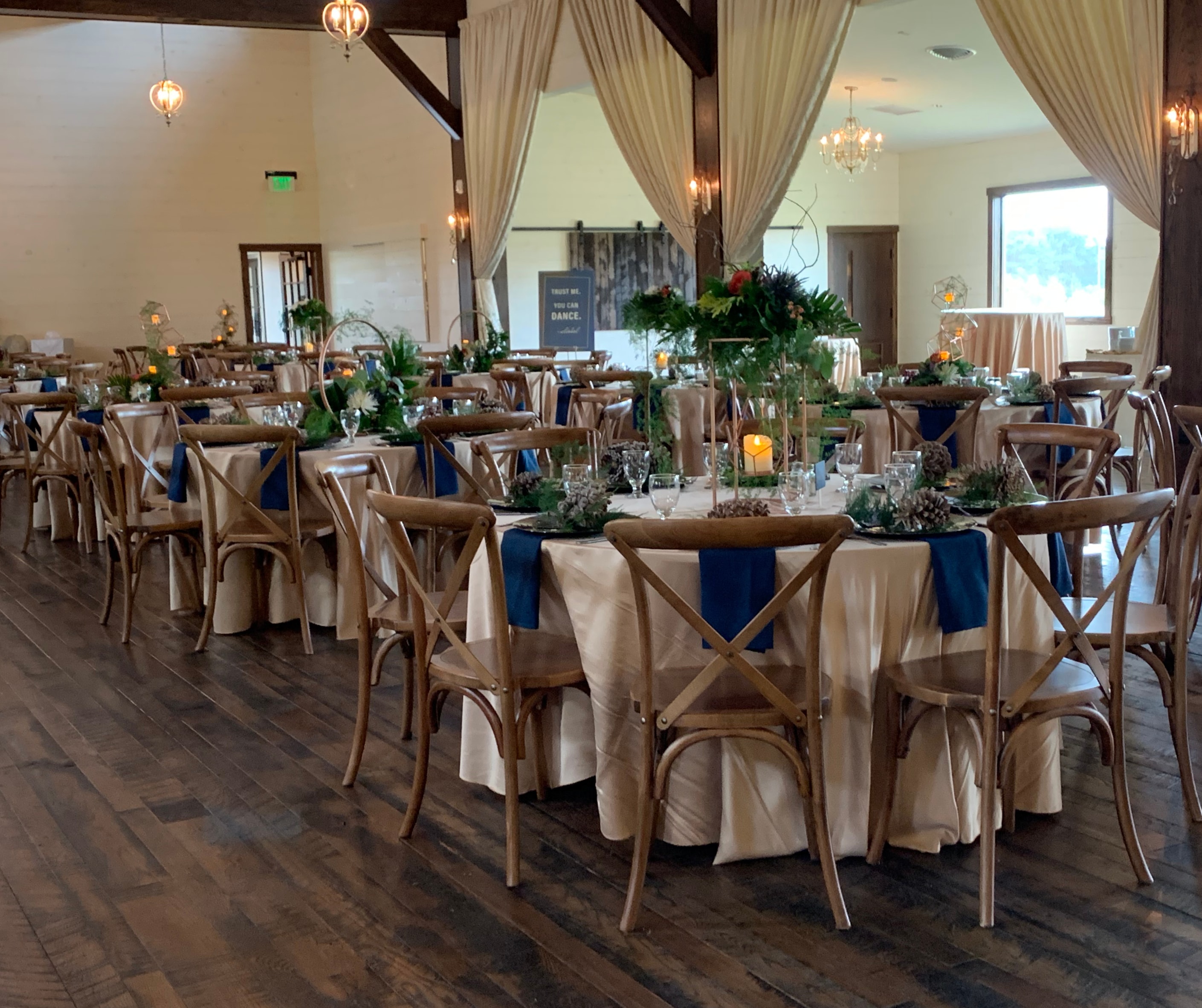 Round guest tables decorated with navy accents, pinecones, greenery, and candles in a rustic venue with wood floors and chand