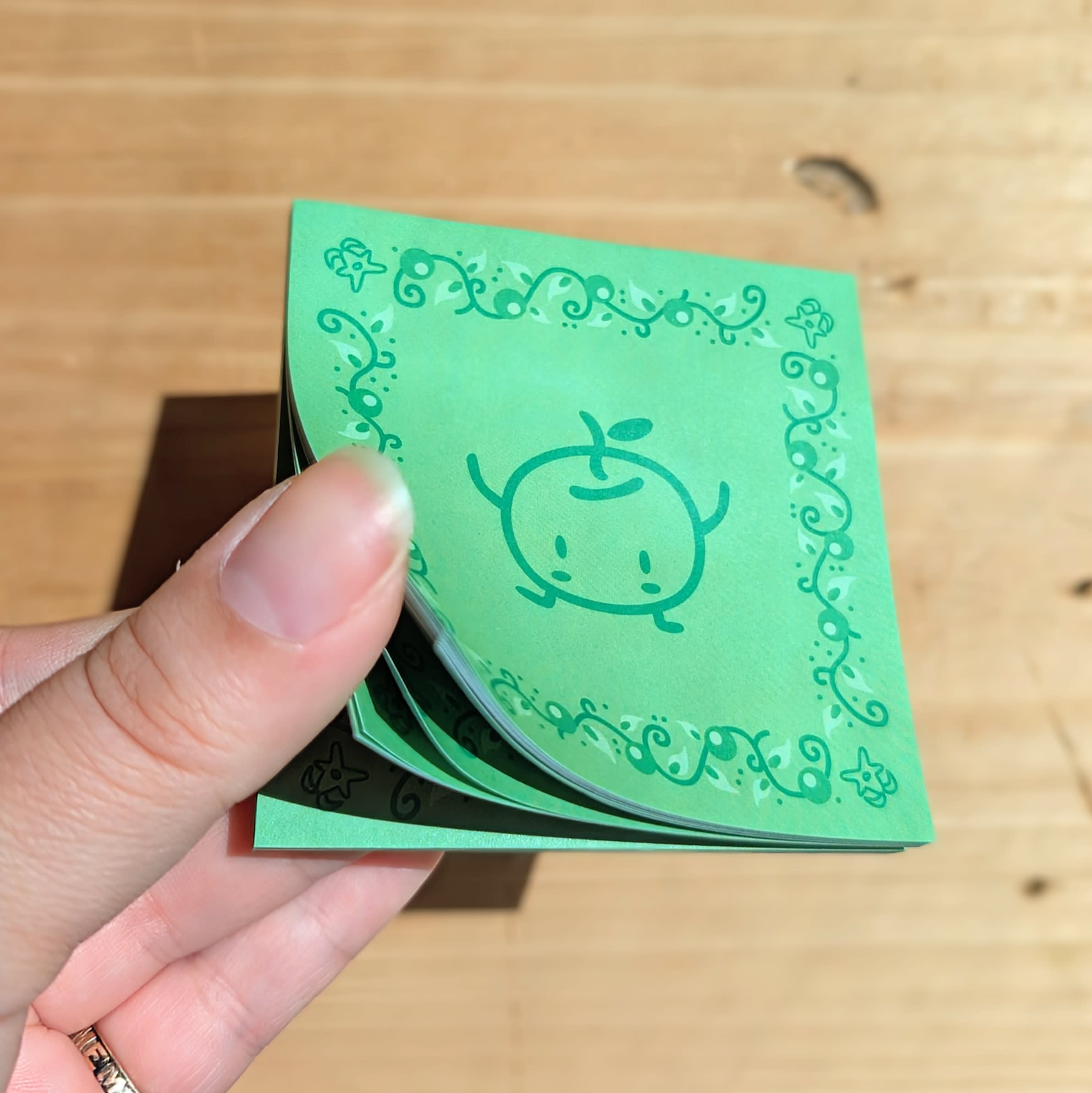 Spring green sticky notepad held in hand with pages splayed. Junimo (forest spirit) art on sheet with leaf border