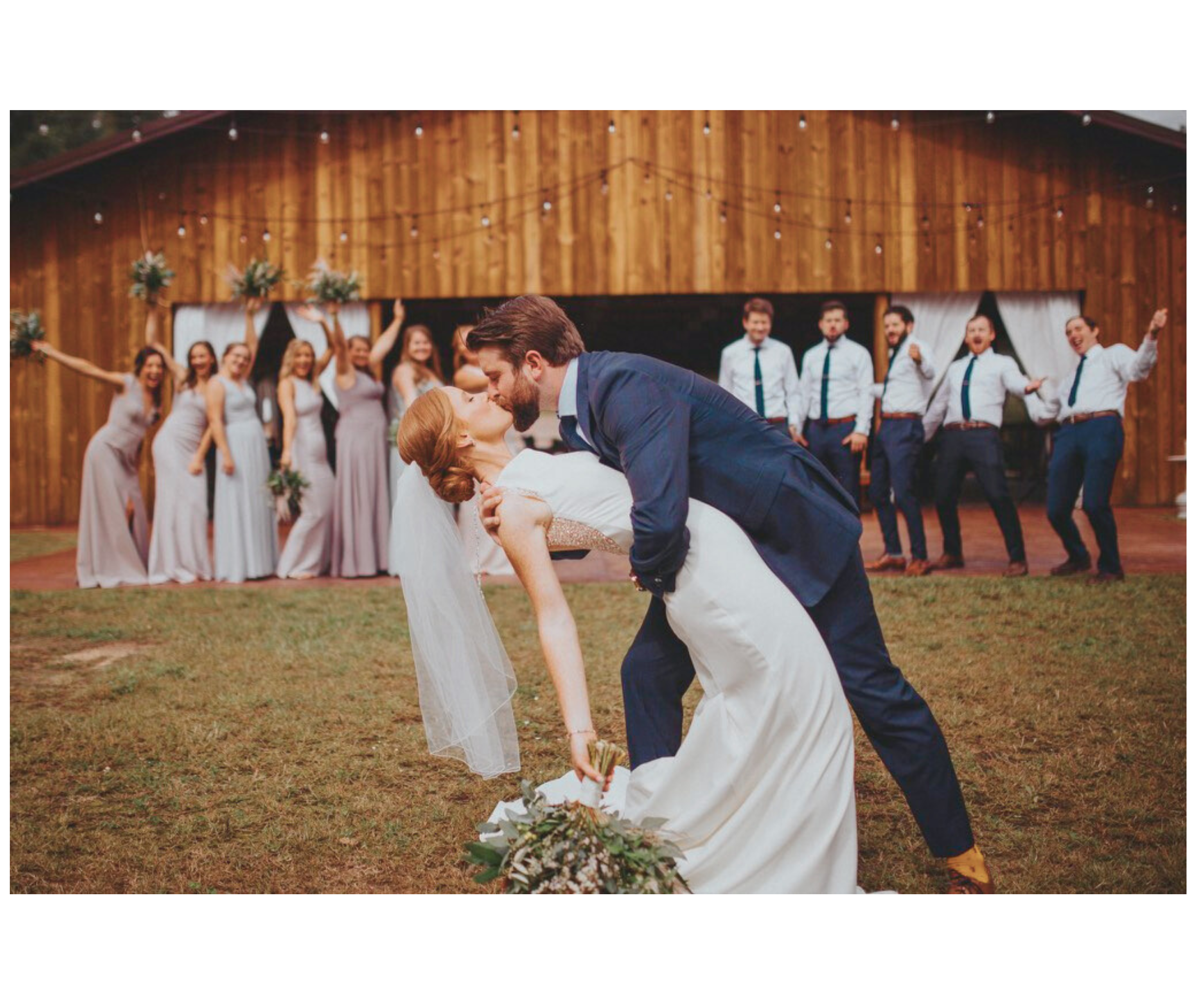 Bride and groom kiss in front of a barn while their wedding party cheers in the background.