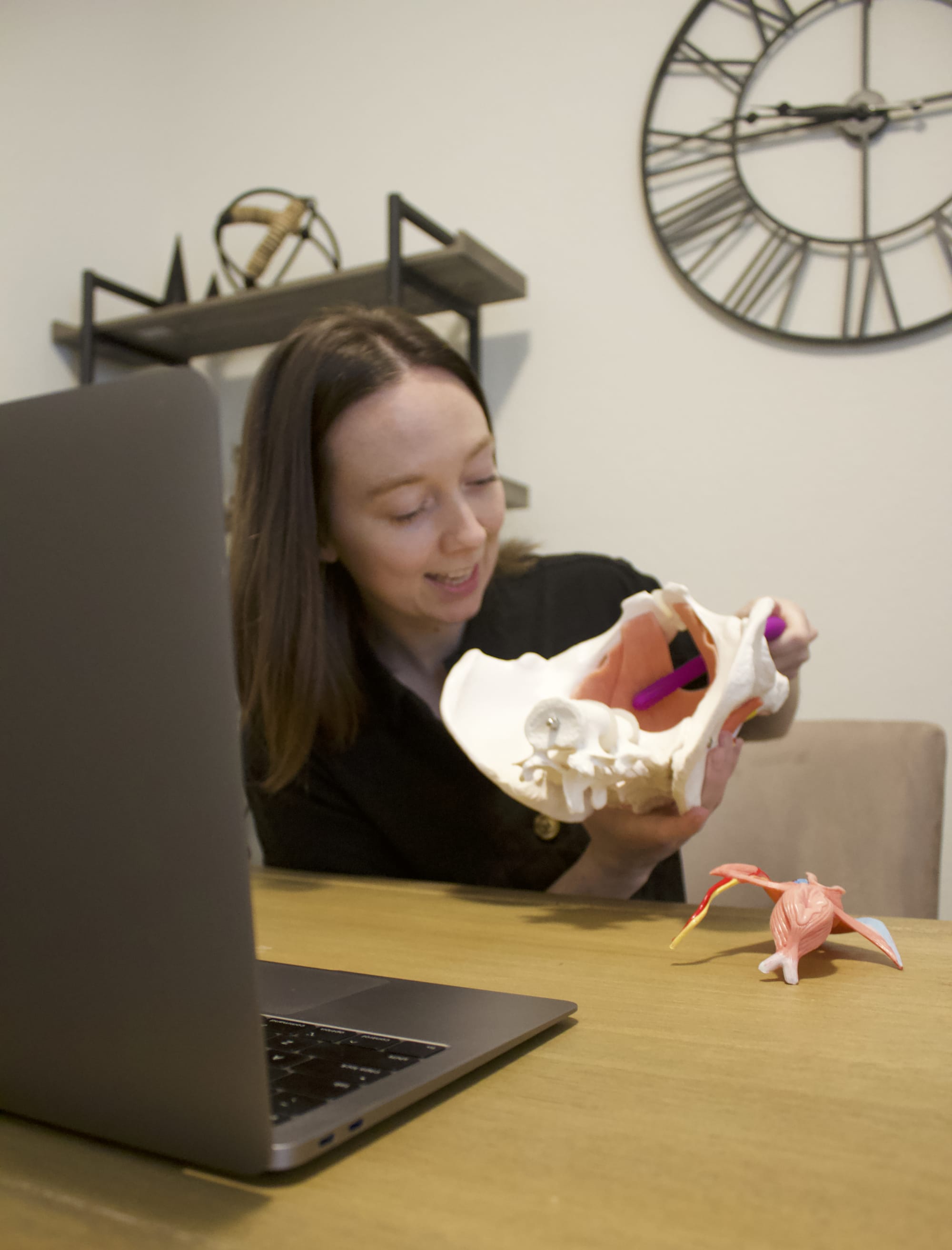 Dr. Ashe sitting at her laptop holding a 3D pelvis model, educating a client how to use a pelvic wand.