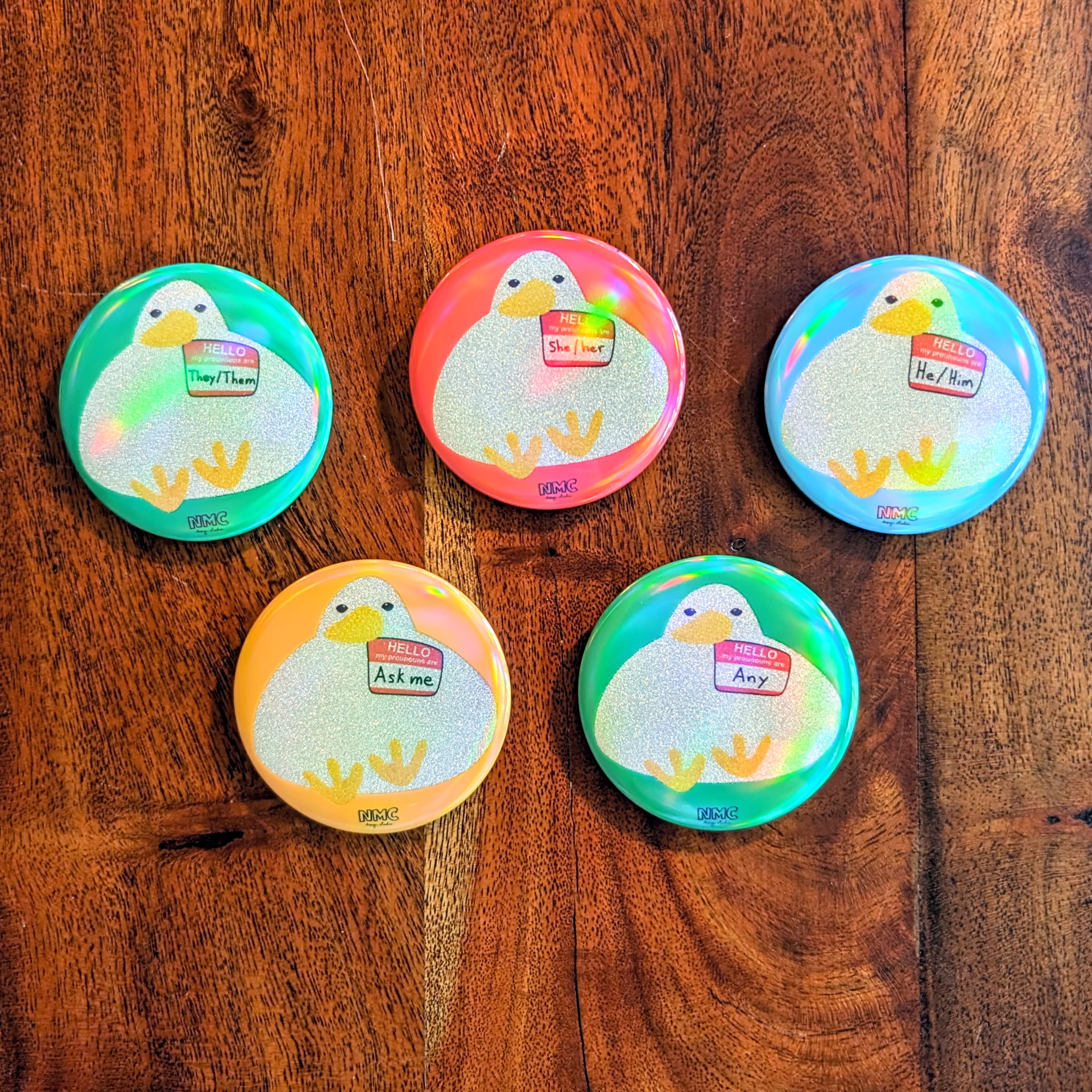 five button pins with glitter and holographic finish. Pins have duck with "name" tag that states different pronouns