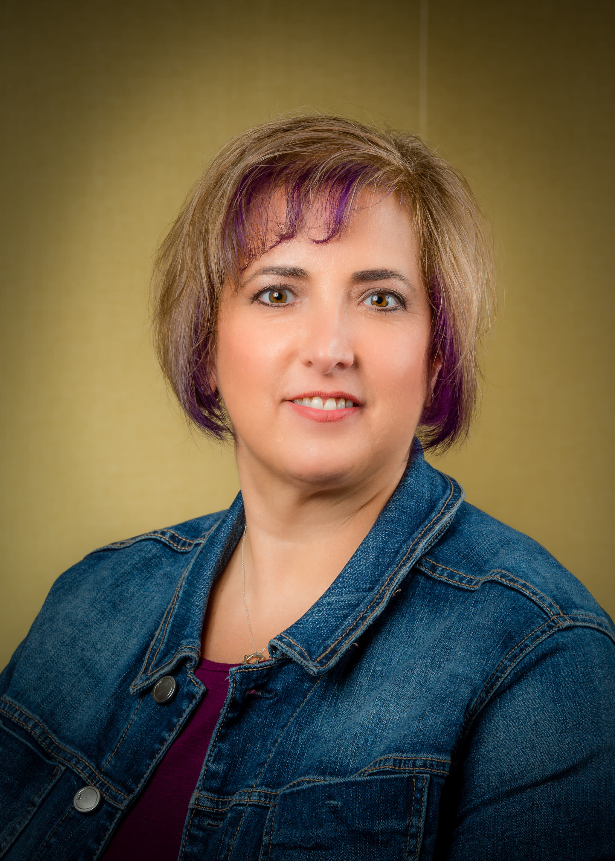 Smiling female with brown hair and blonde and purple highlights wearing a burgundy shirt with jean jacket